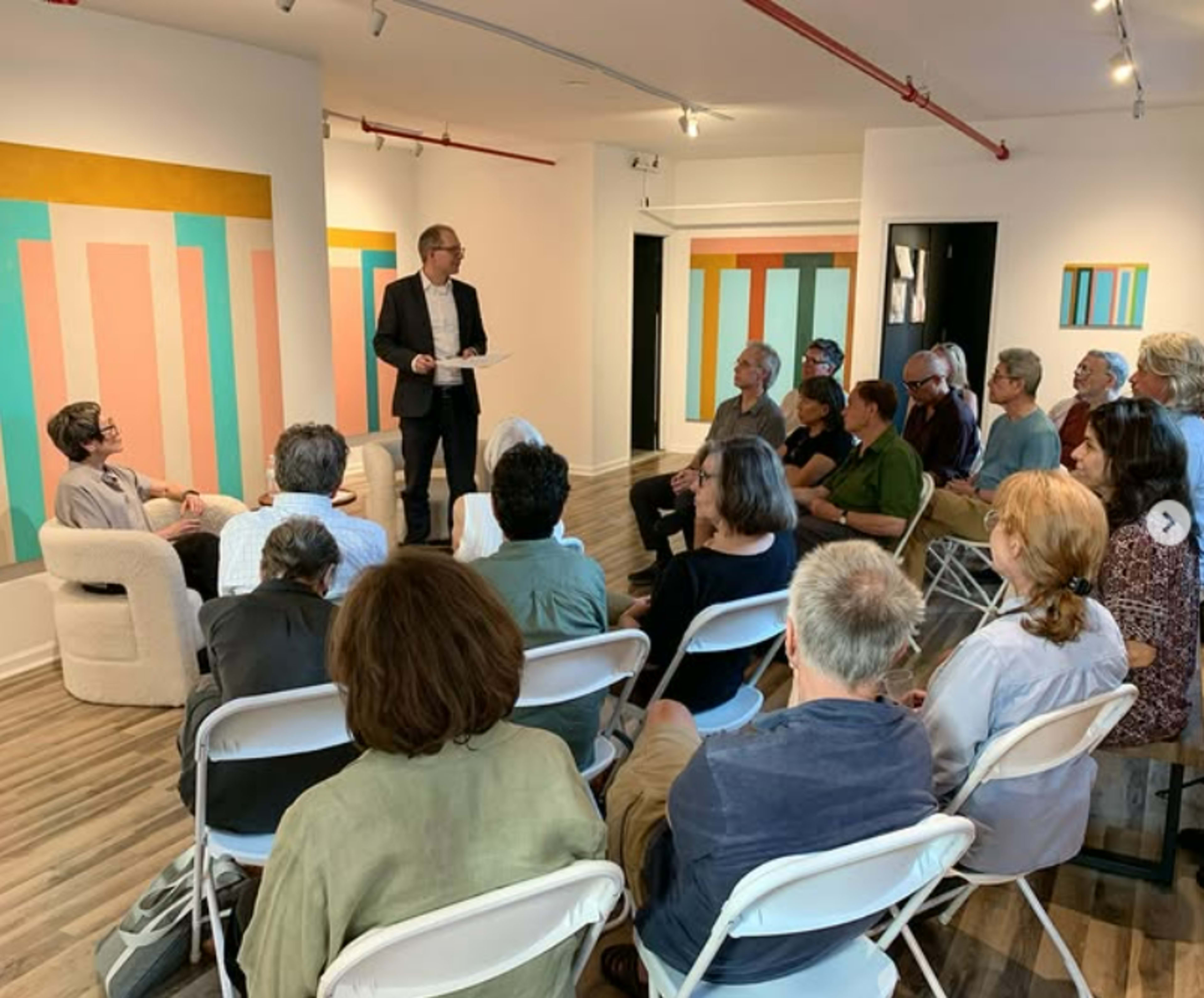 A speaker stands in front of an audience seated in folding chairs, with colorful artwork displayed on the walls of the gallery.