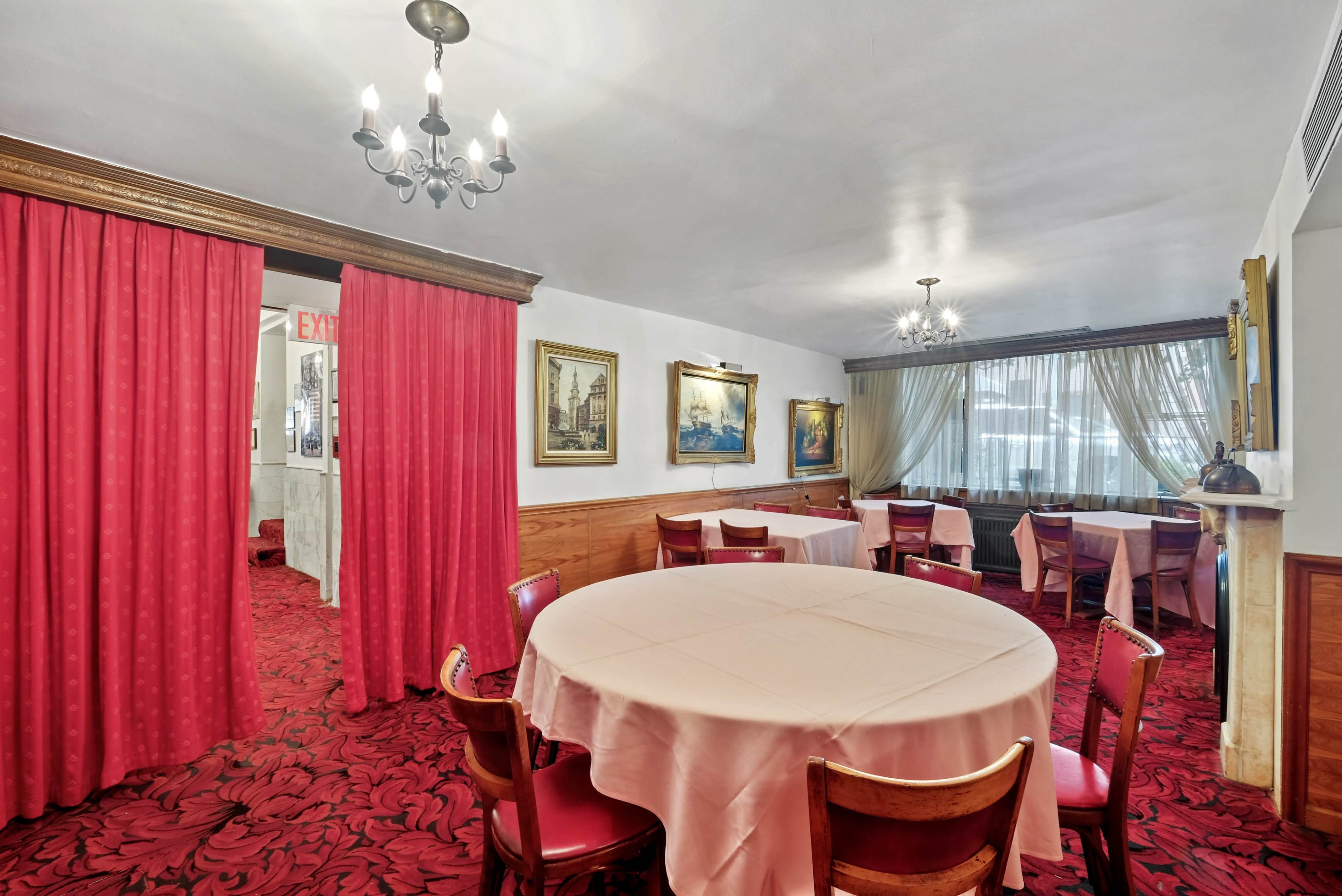 A dining area with round tables covered in pink tablecloths, red carpeting, and framed paintings on the walls.