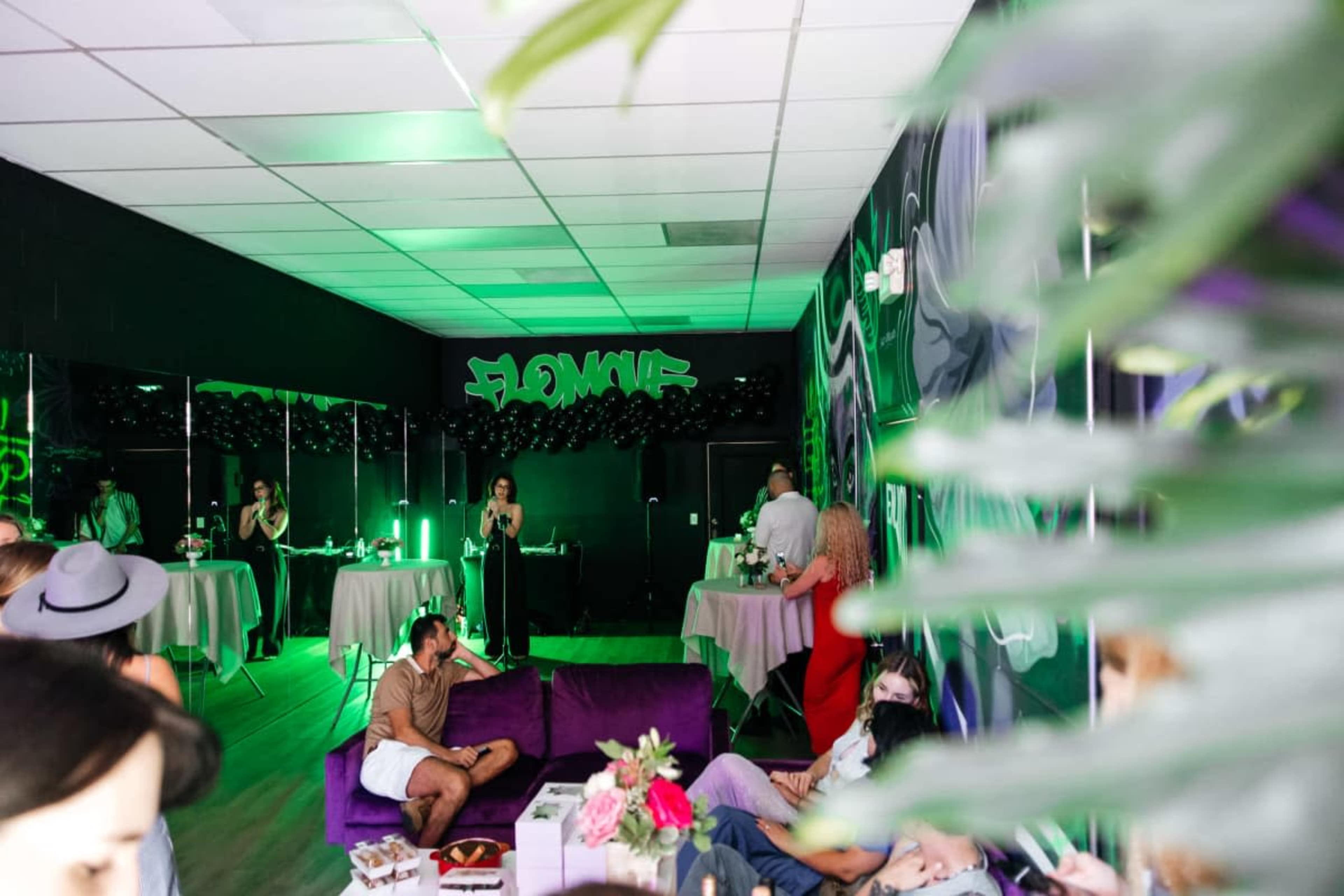 A vibrant indoor event space features people socializing around tables, with a colorful graffiti backdrop and green lighting.