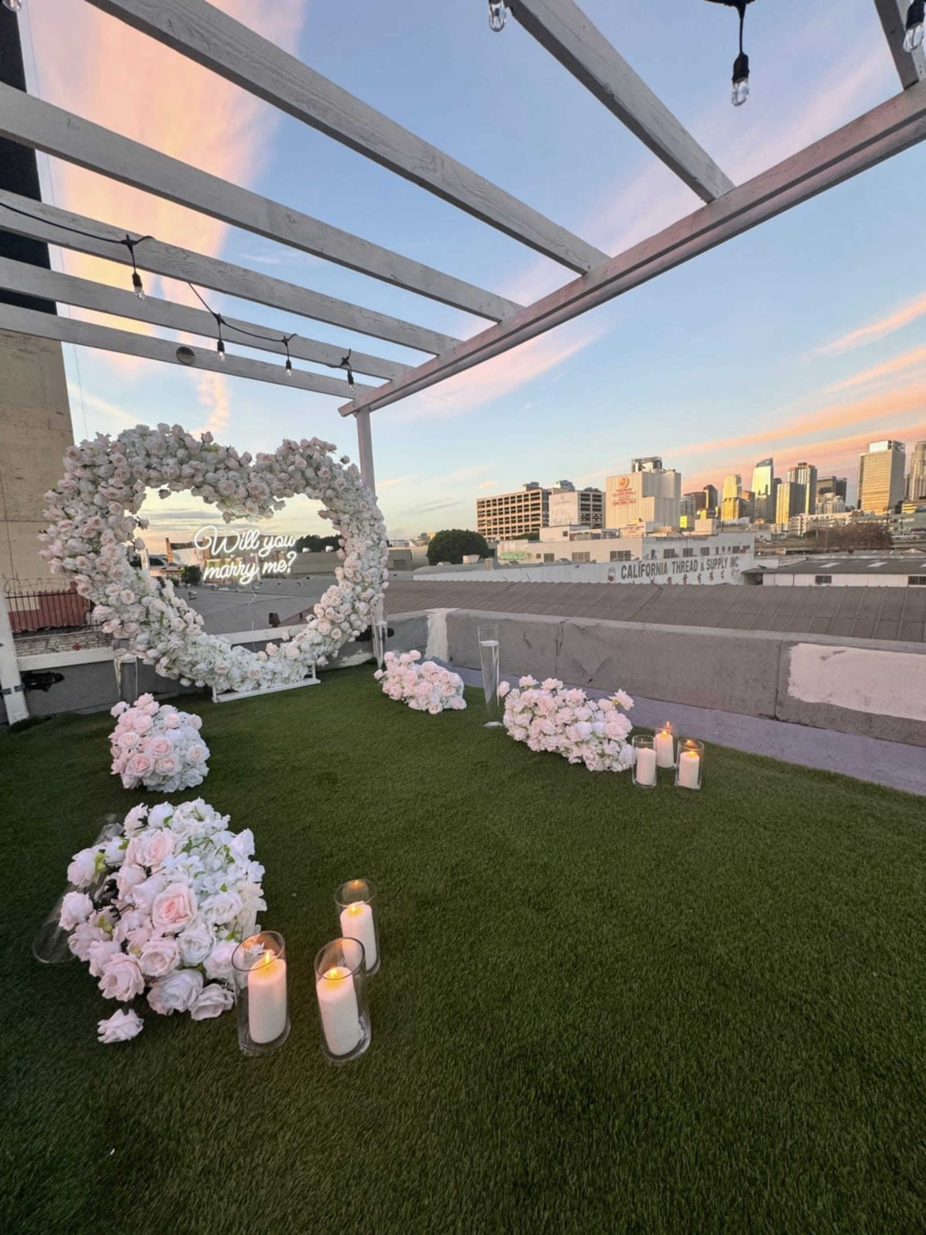 A rooftop space adorned with a floral heart display and candles, overlooking a city skyline at sunset.