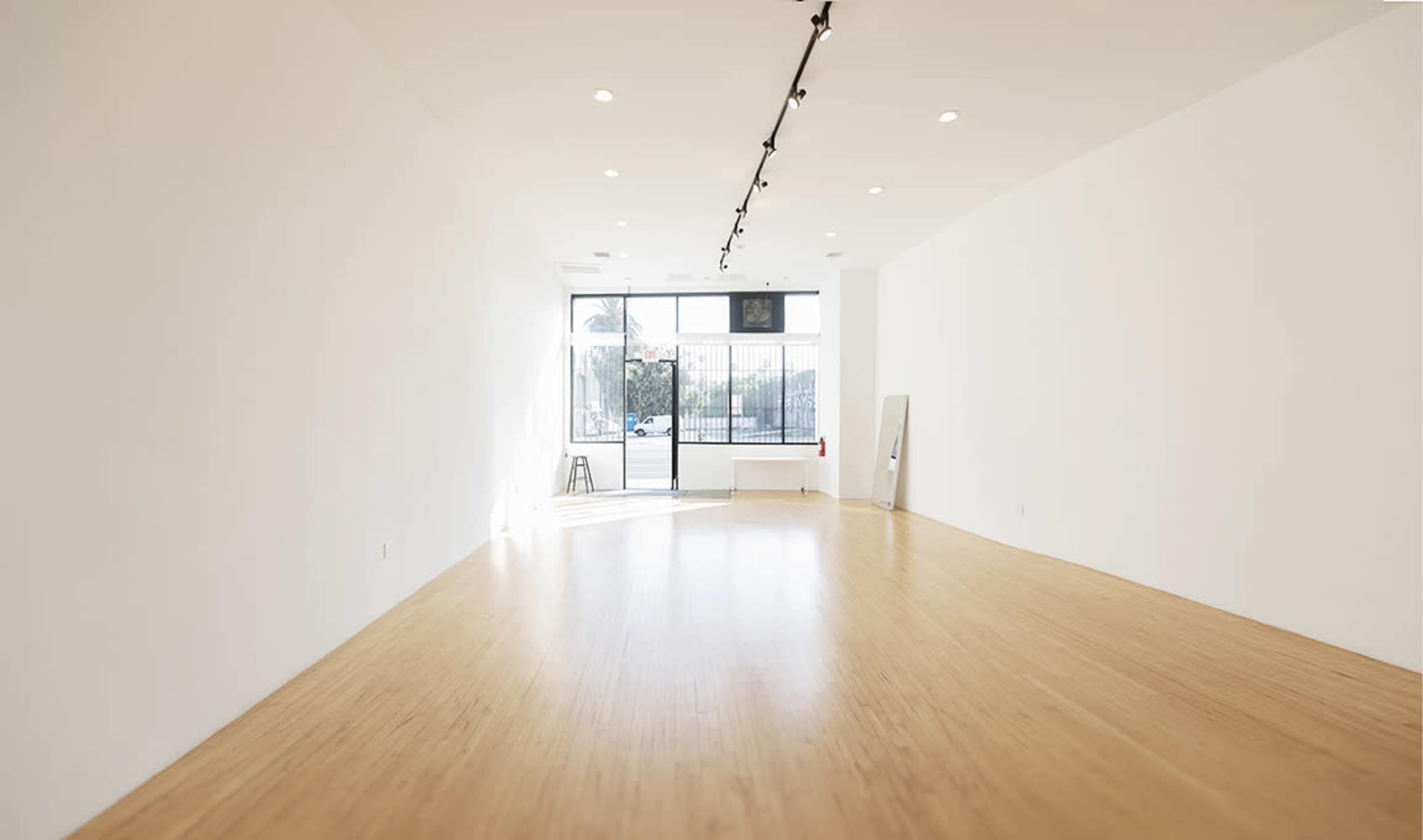 The image shows a spacious, empty room with bamboo flooring and large windows that let in natural light.
