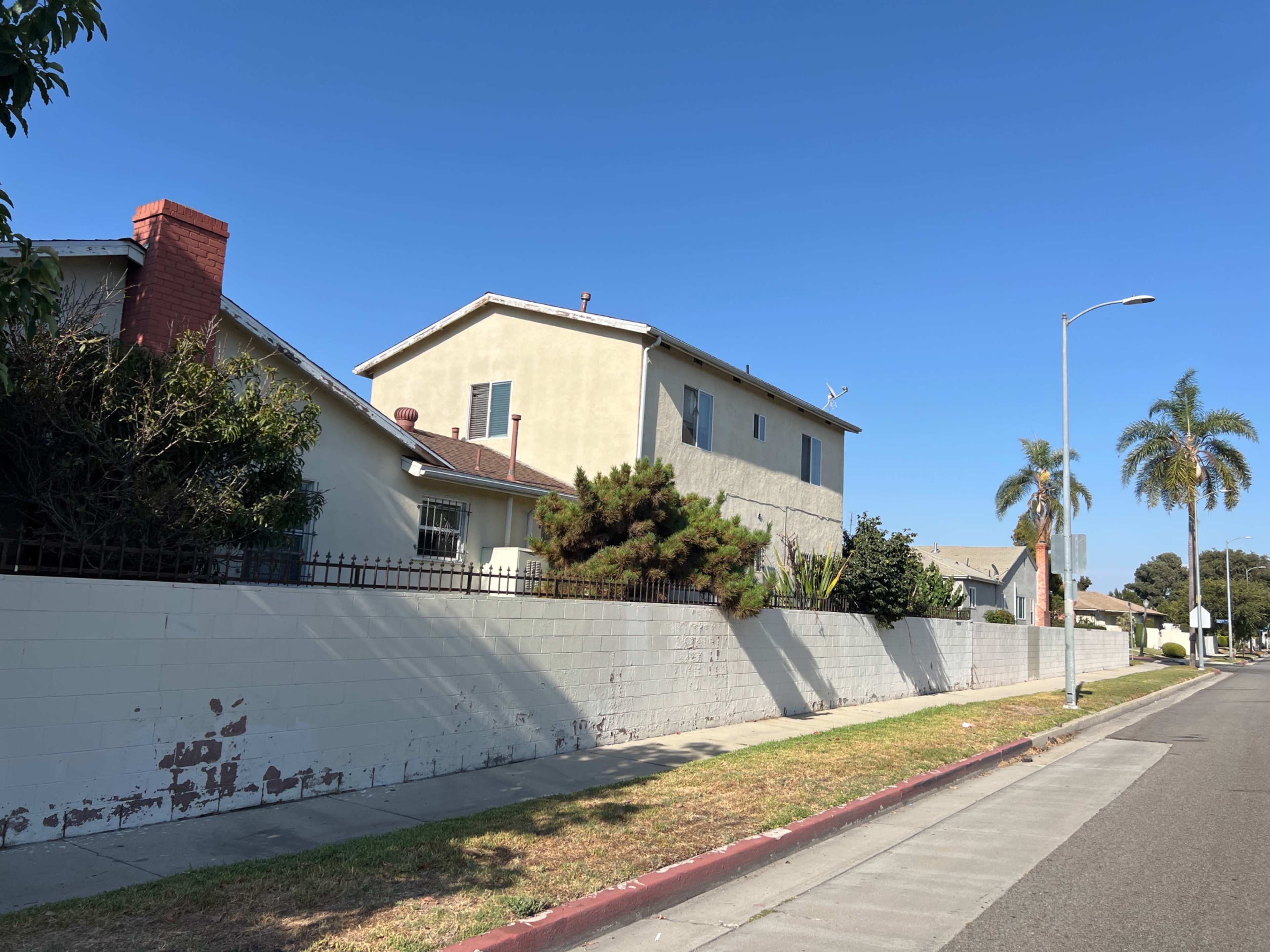 A two-story house with a pale exterior stands next to a low white wall lined with palm trees along a quiet street.