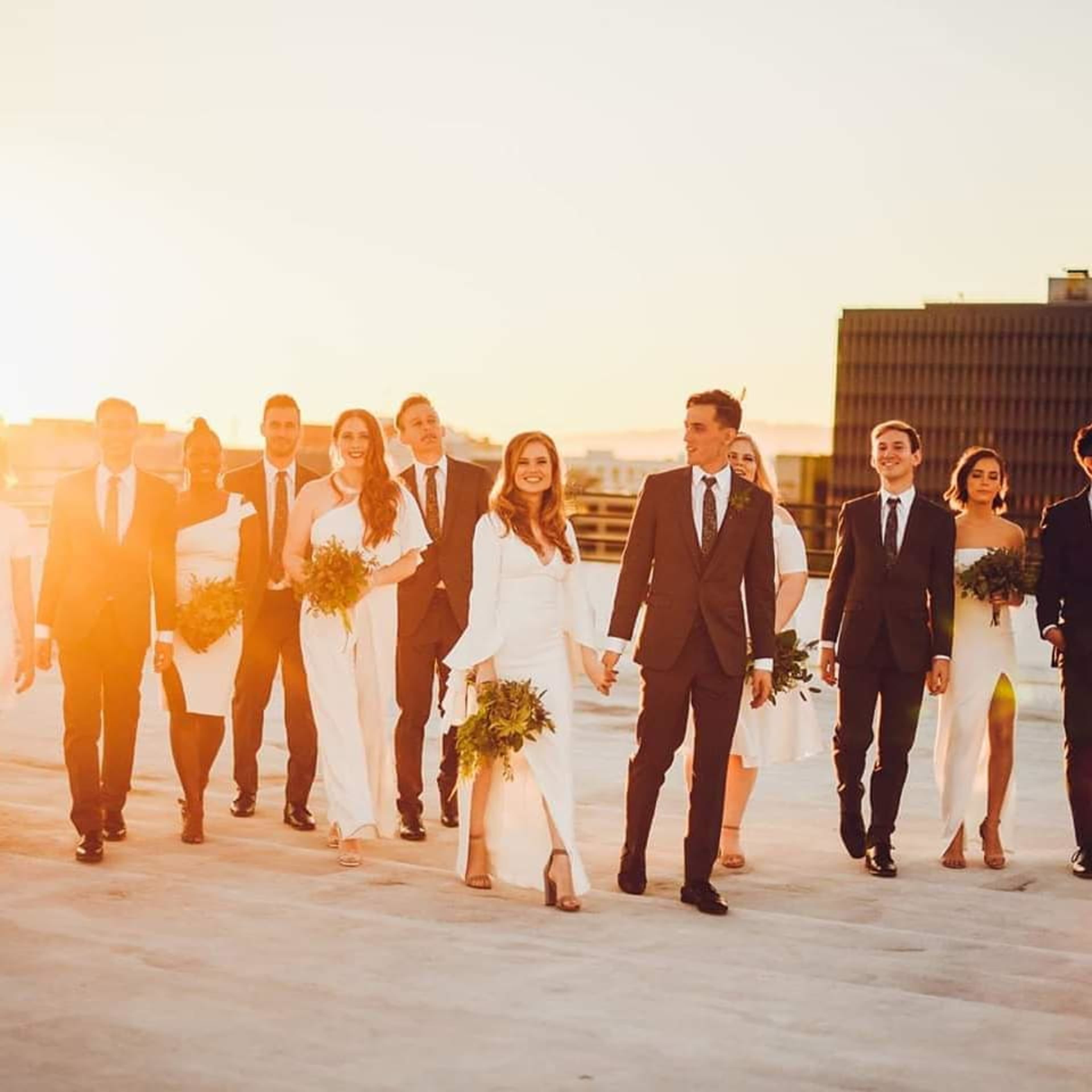 A wedding party in formal attire walks together at sunset, with the bride and groom holding hands at the front.