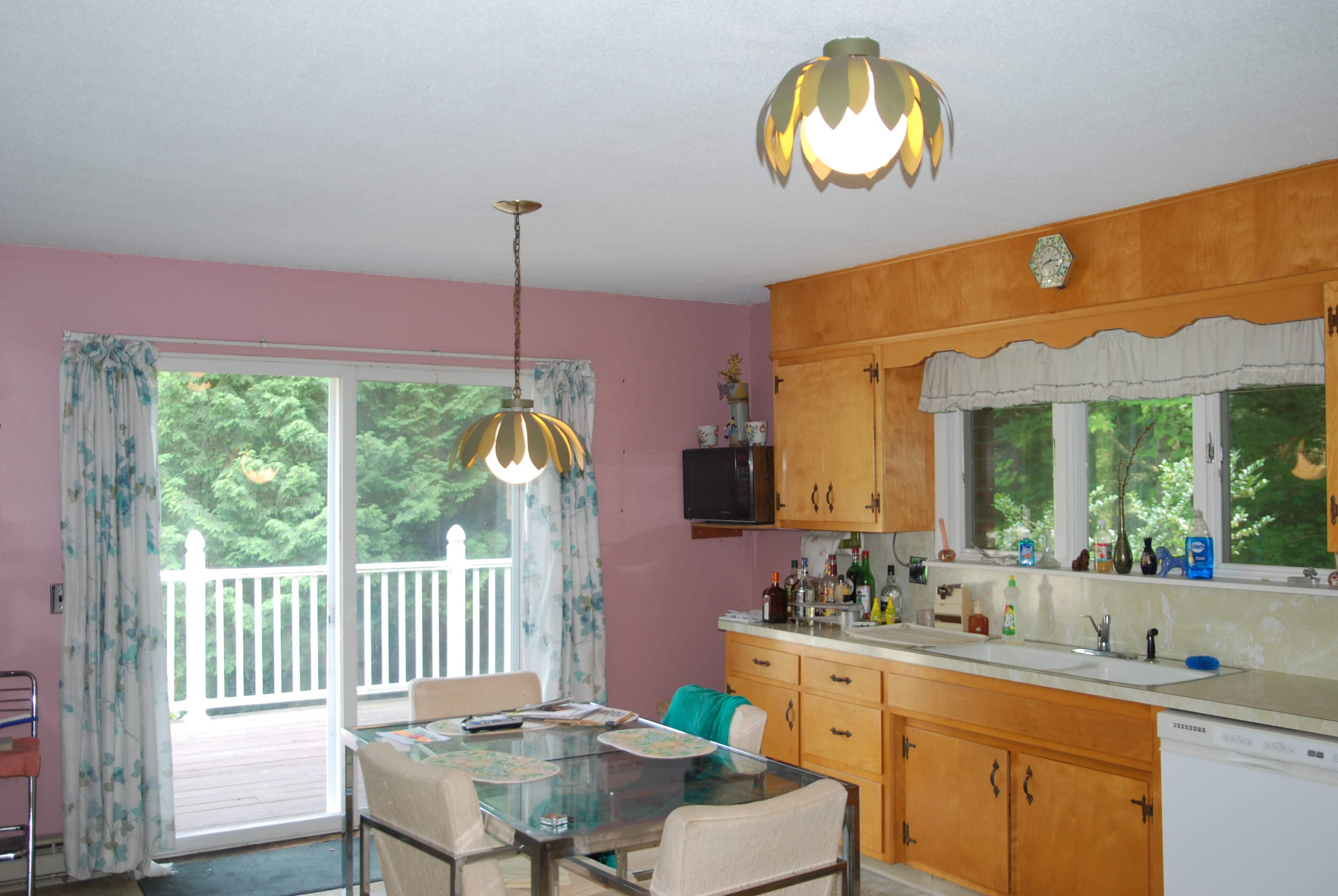 The image shows a kitchen with light wood cabinetry, a glass dining table with four chairs, and a sliding glass door leading to an outdoor deck surrounded by greenery.
