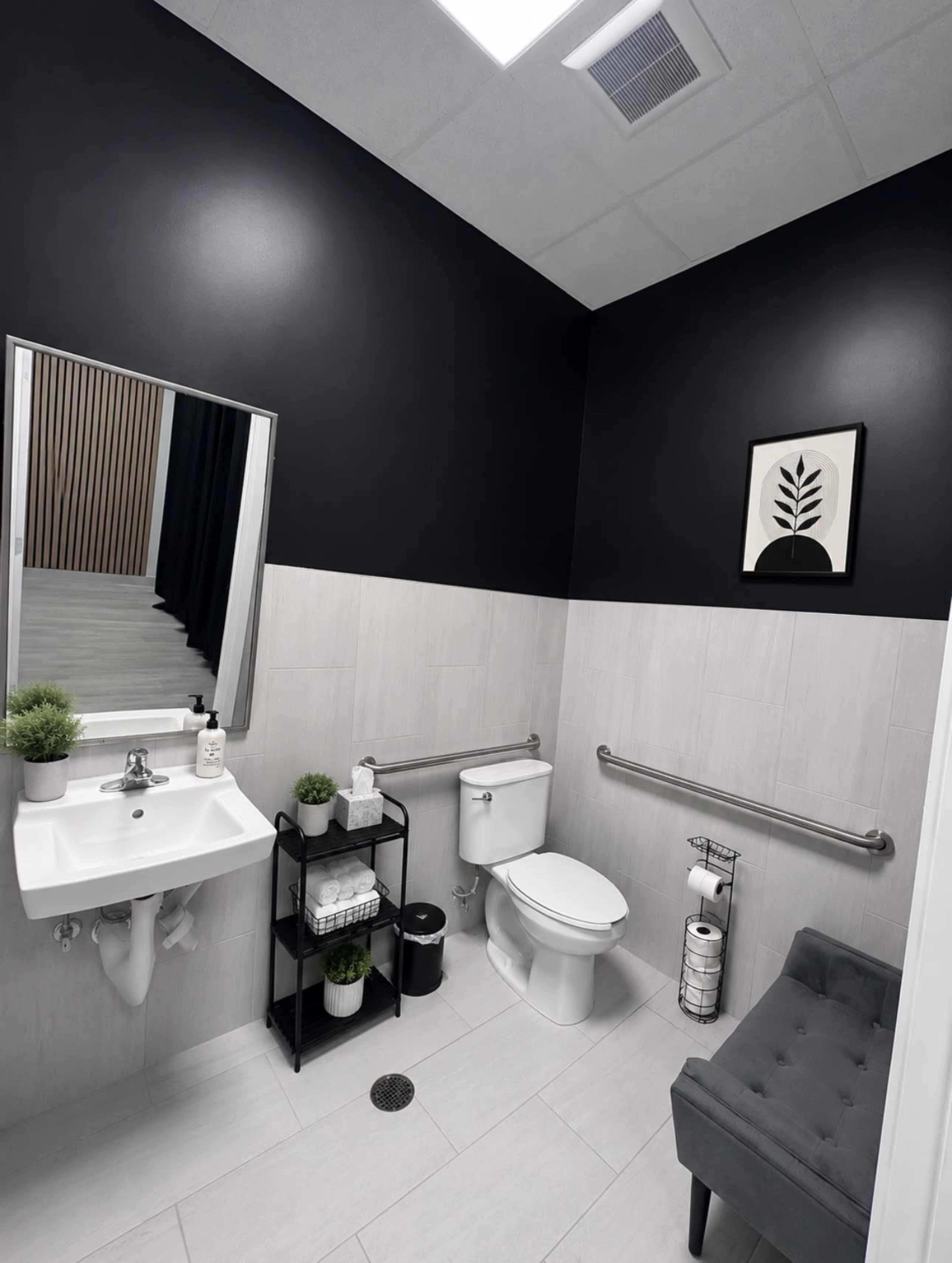 The image shows a modern bathroom with dark walls, a large mirror, a toilet, a small sink, and a gray bench, all arranged with plants and grooming supplies.