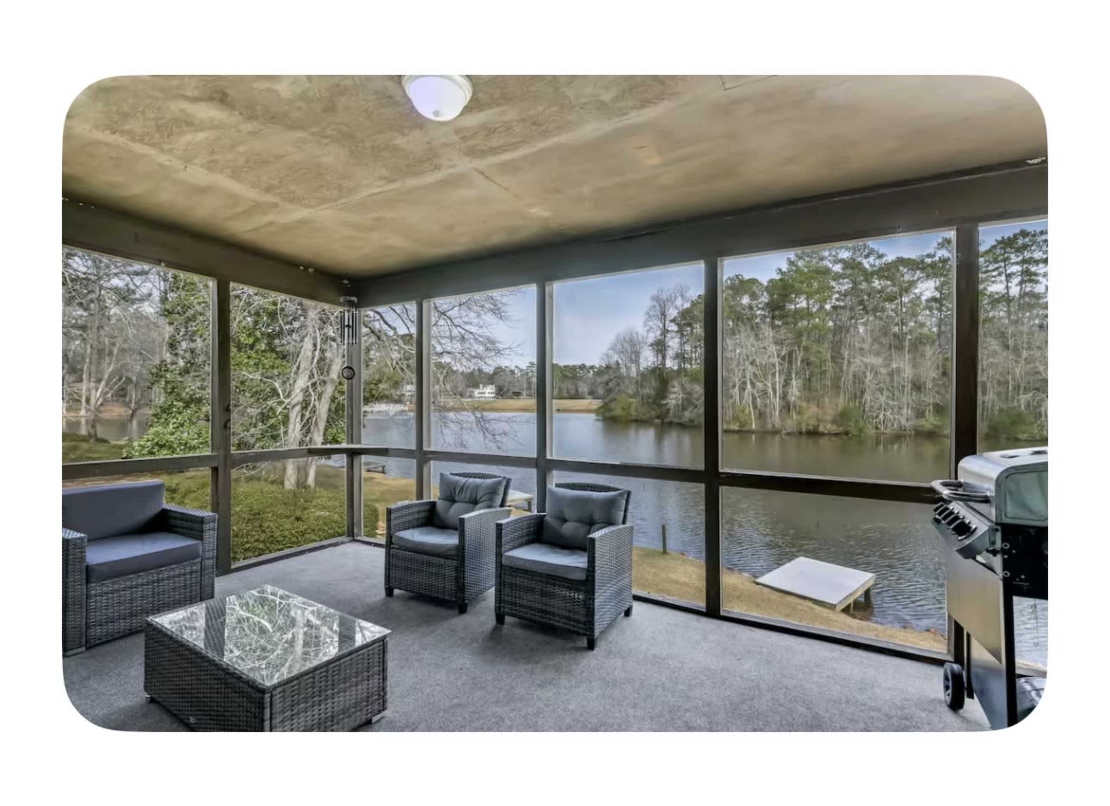 The image shows a screened-in patio overlooking a calm lake, furnished with a glass-topped table and wicker chairs.