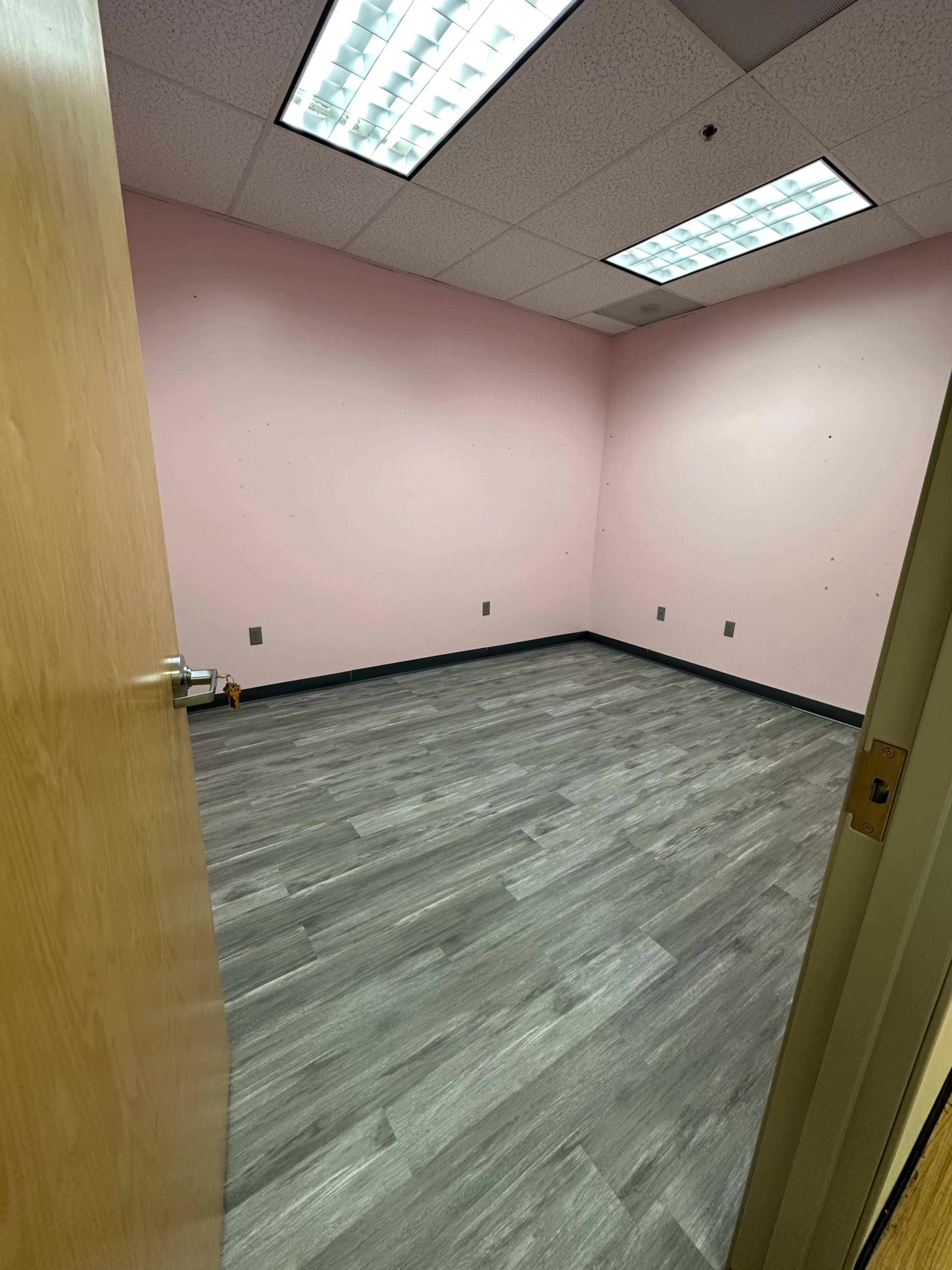 An empty room with pink walls and gray laminate flooring, featuring fluorescent ceiling lights and electrical outlets on the walls.