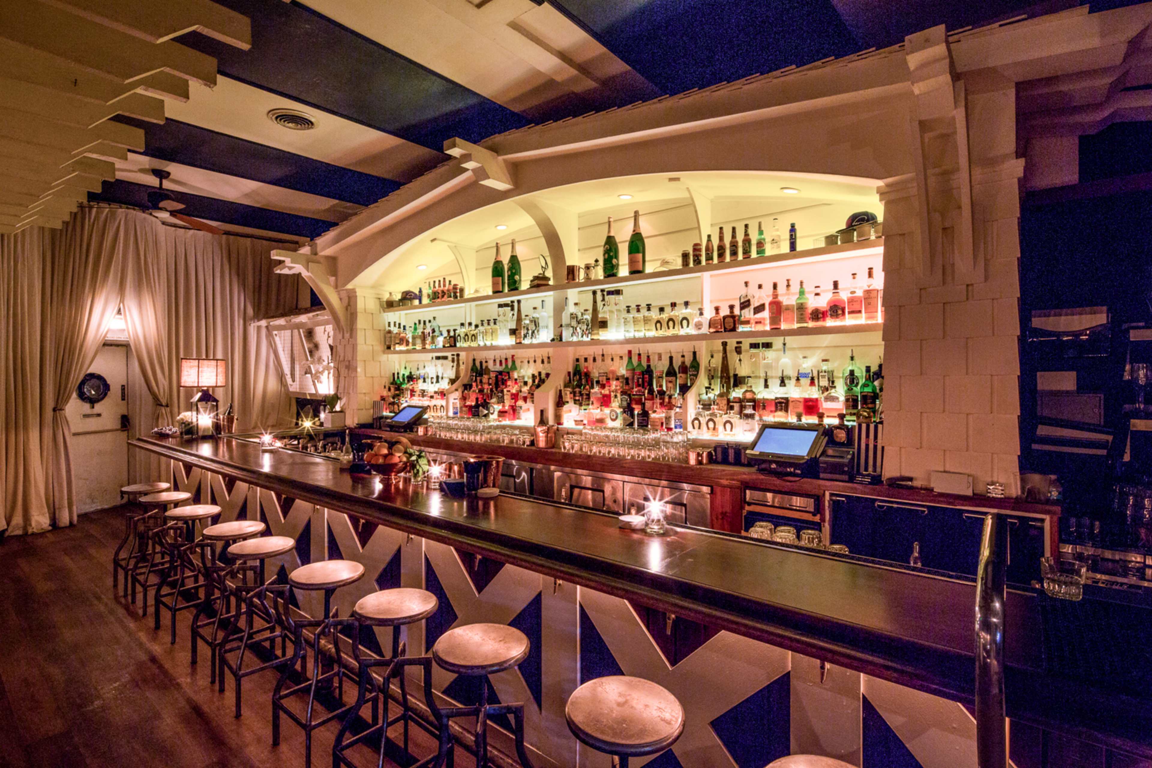 A well-lit bar with a wide selection of bottles displayed on shelves behind the counter and several stools in front.