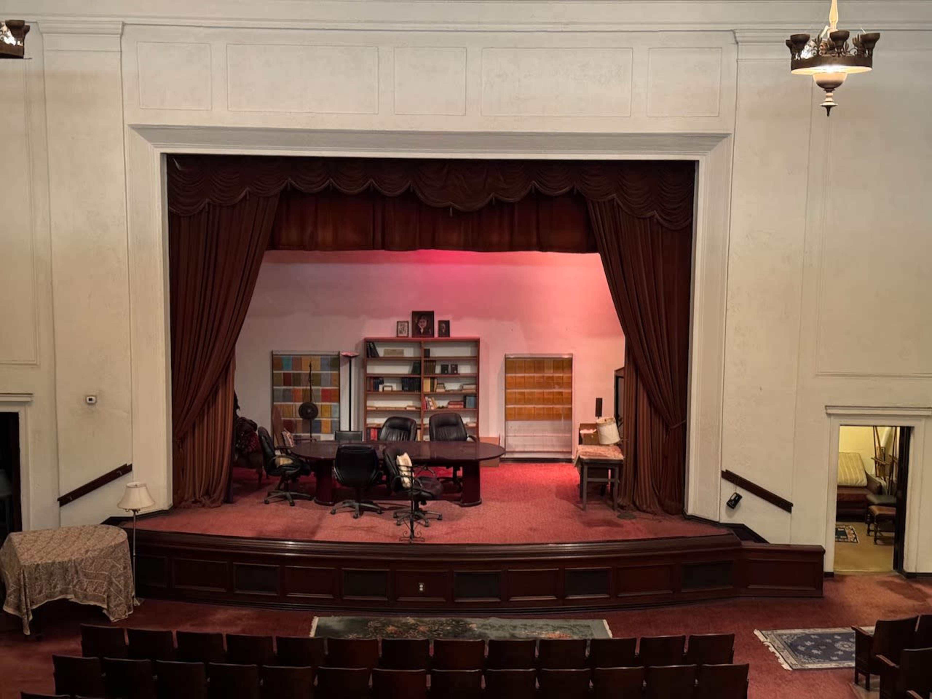The image shows a stage setup with a decorated backdrop featuring an office scene, complete with a large desk, chairs, and shelves, all illuminated by soft lighting.