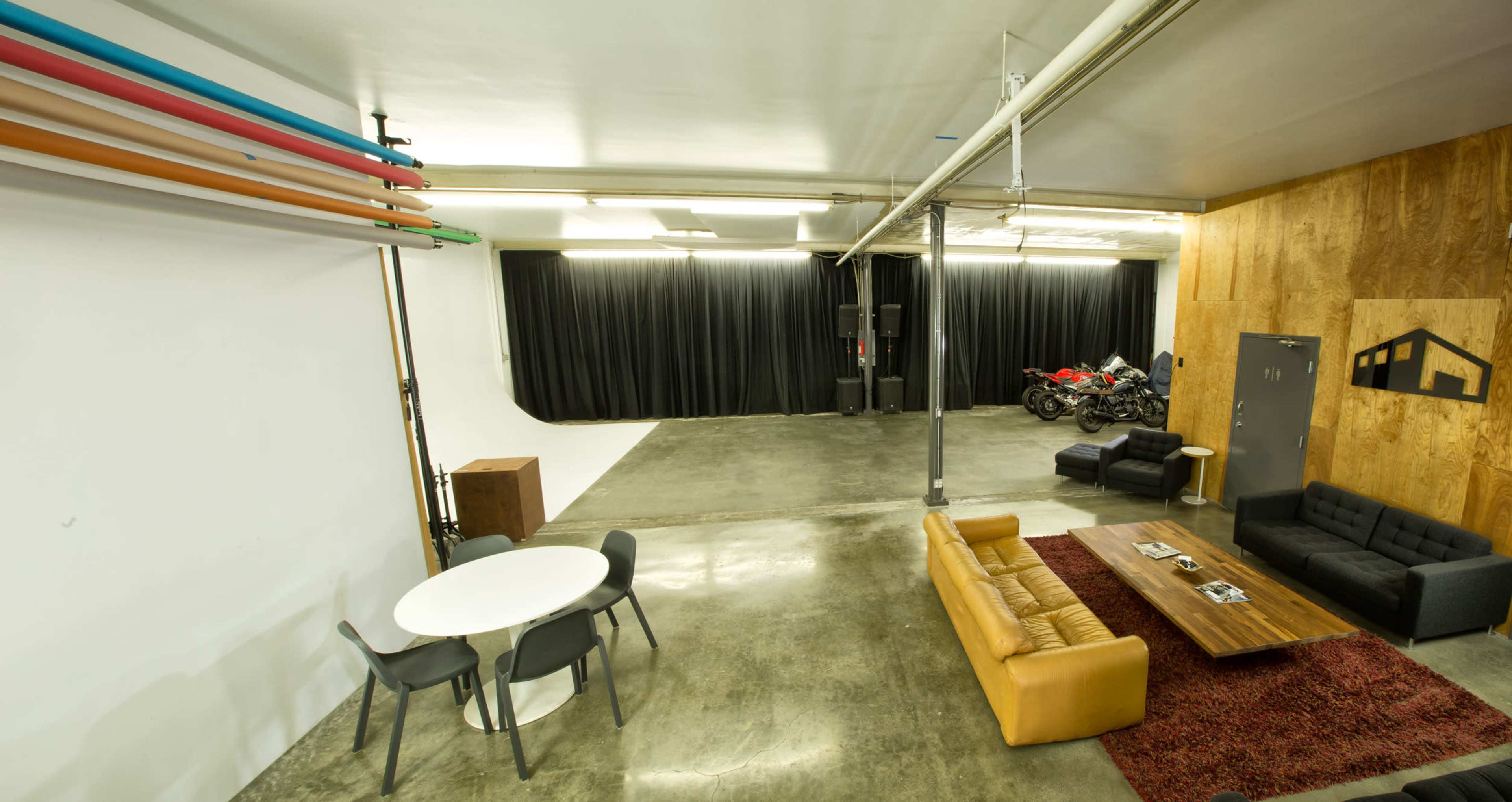 The image shows a spacious, industrial-style studio with a seating area, a dining table, and a backdrop with black curtains, along with some motorcycles parked against one wall.