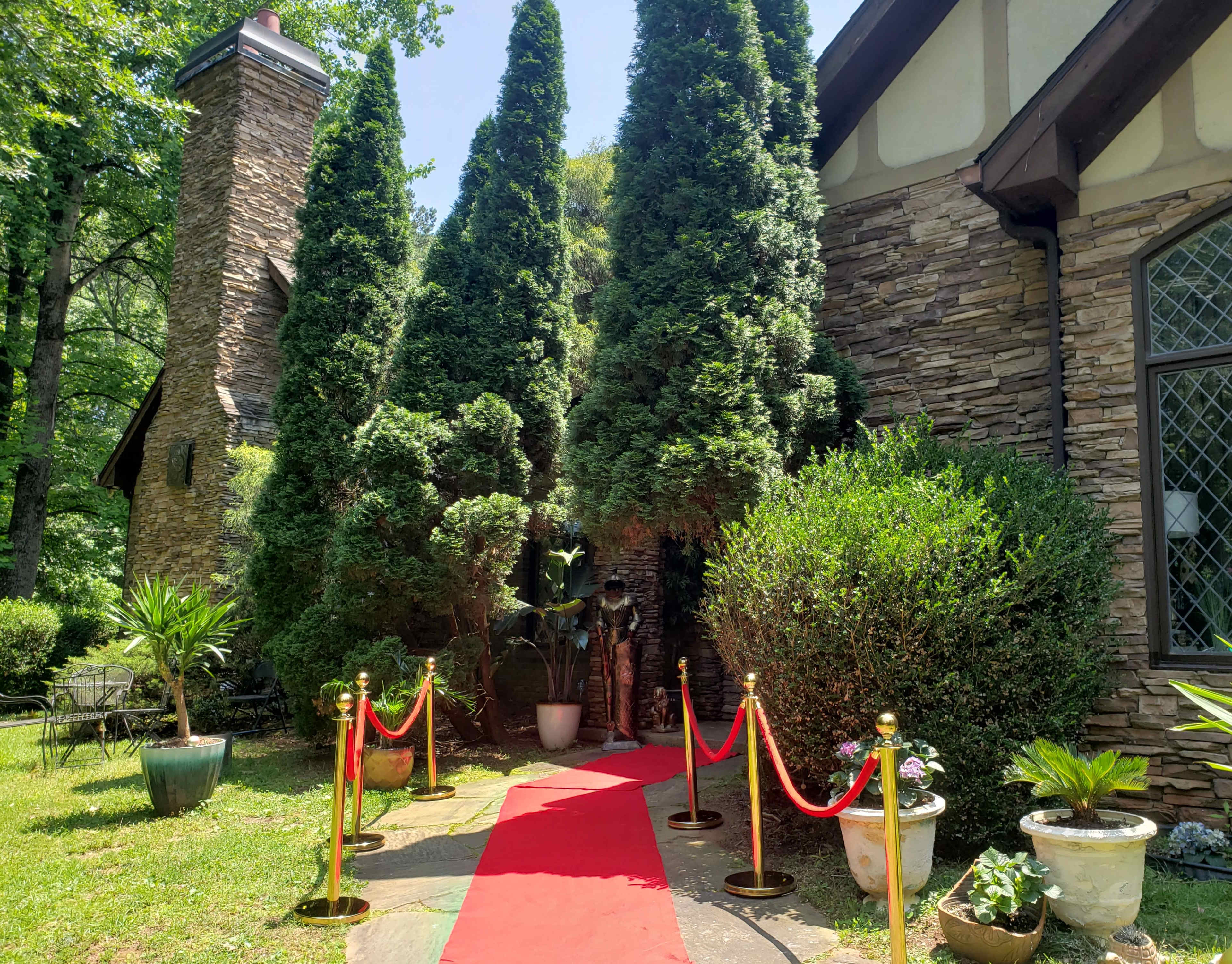 A red carpet leads to a stone house surrounded by tall, lush conifer trees and neatly arranged potted plants.