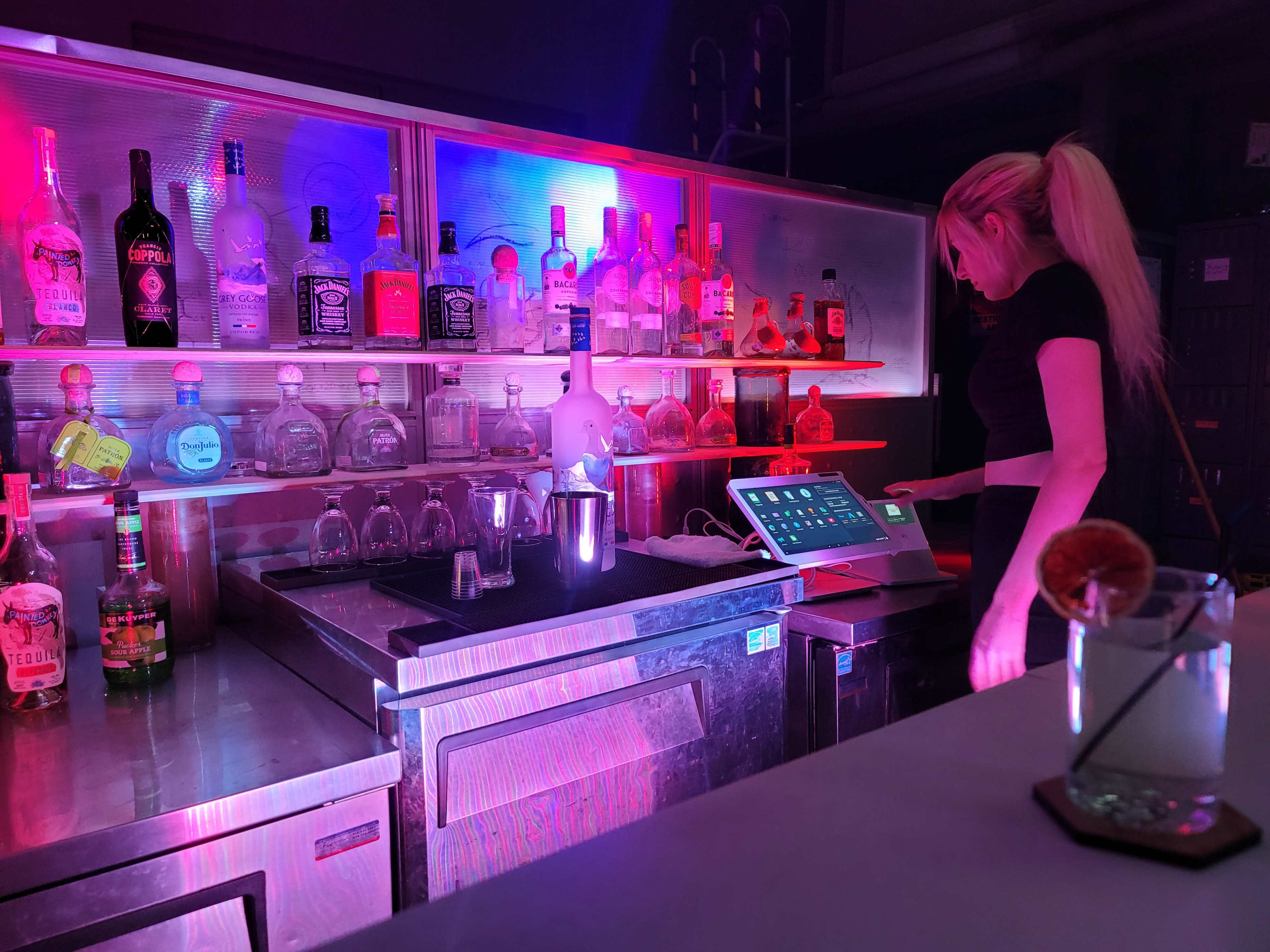 A bartender stands at a brightly lit bar with various bottles and a tablet display.