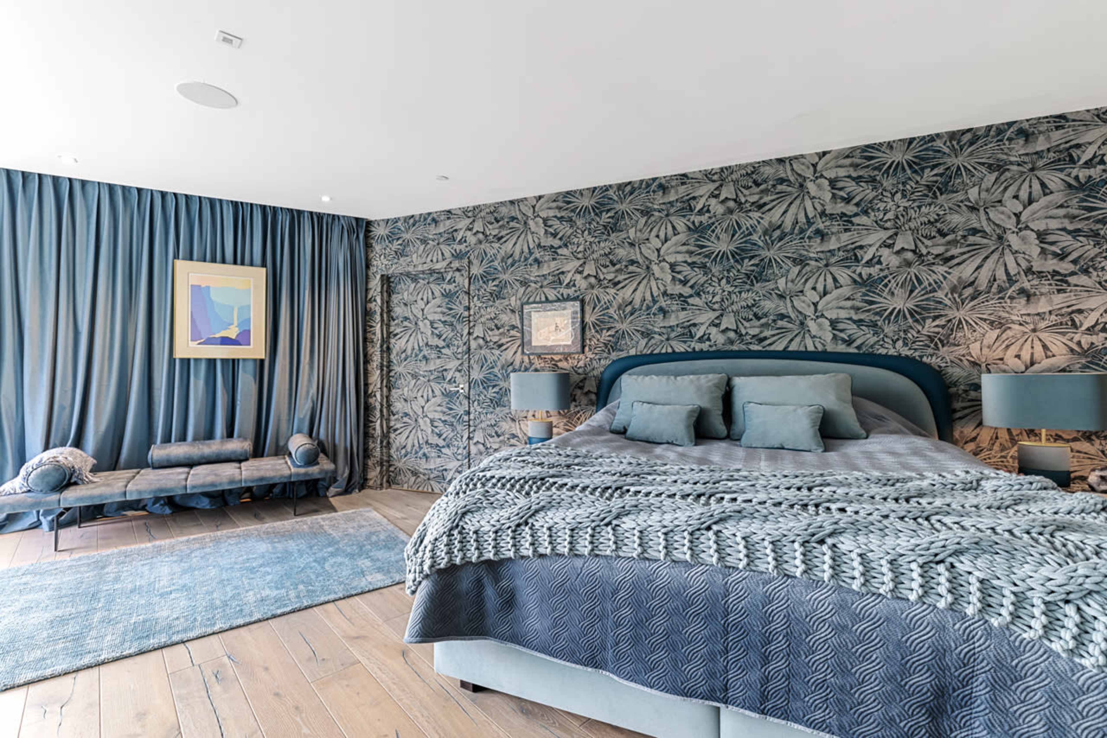 The image shows a modern bedroom featuring a bed with a patterned throw, blue decor, and a large window draped with blue curtains.