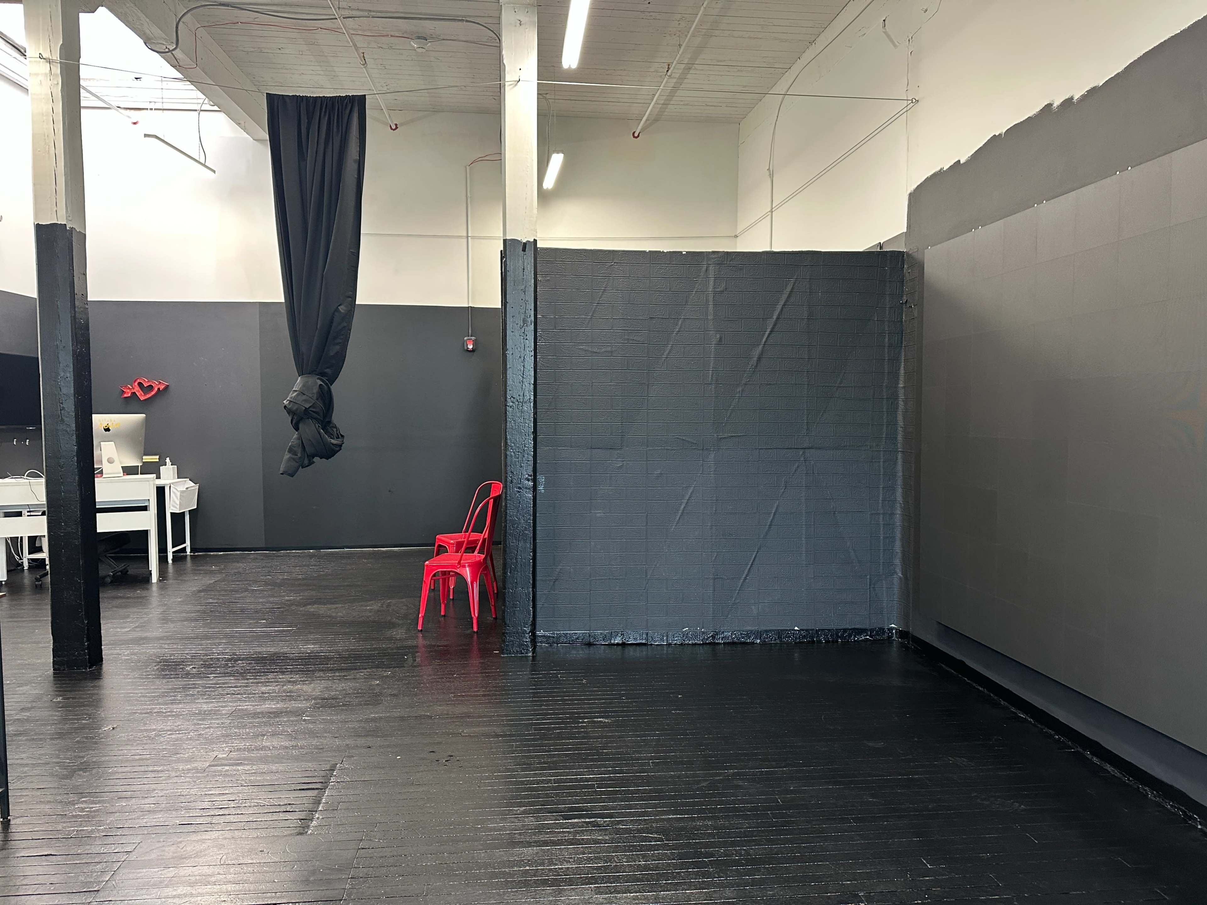 The image shows an empty room with dark gray walls, a black fabric draped from the ceiling, a red chair, and a small work area with a desk in the background.