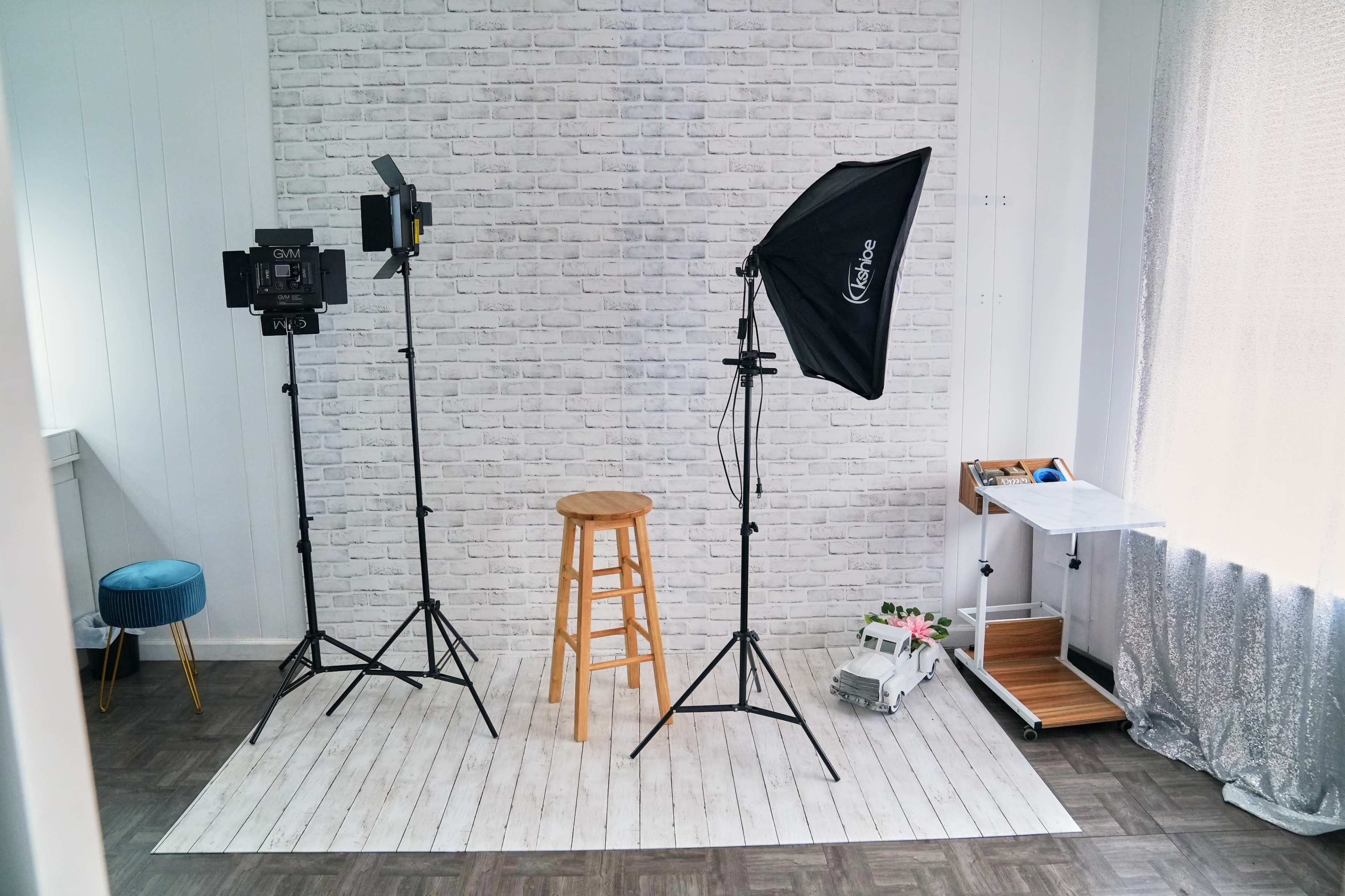 The image shows a photography studio setup featuring a stool, lighting equipment, and a backdrop against a white brick wall.