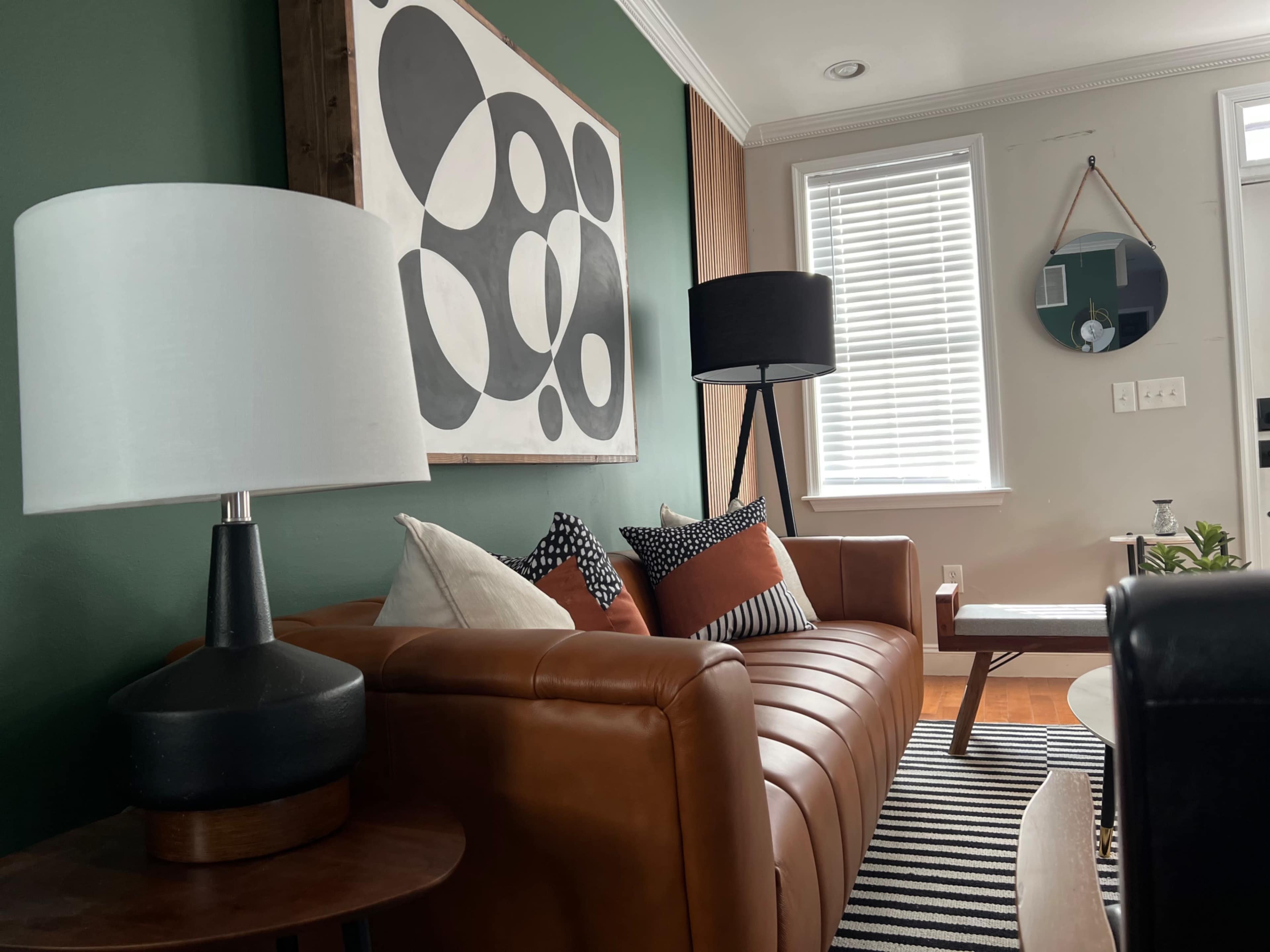 The scene depicts a cozy living room featuring a brown leather sofa with various patterned pillows, a circular coffee table, and modern light fixtures against a green wall adorned with abstract art.