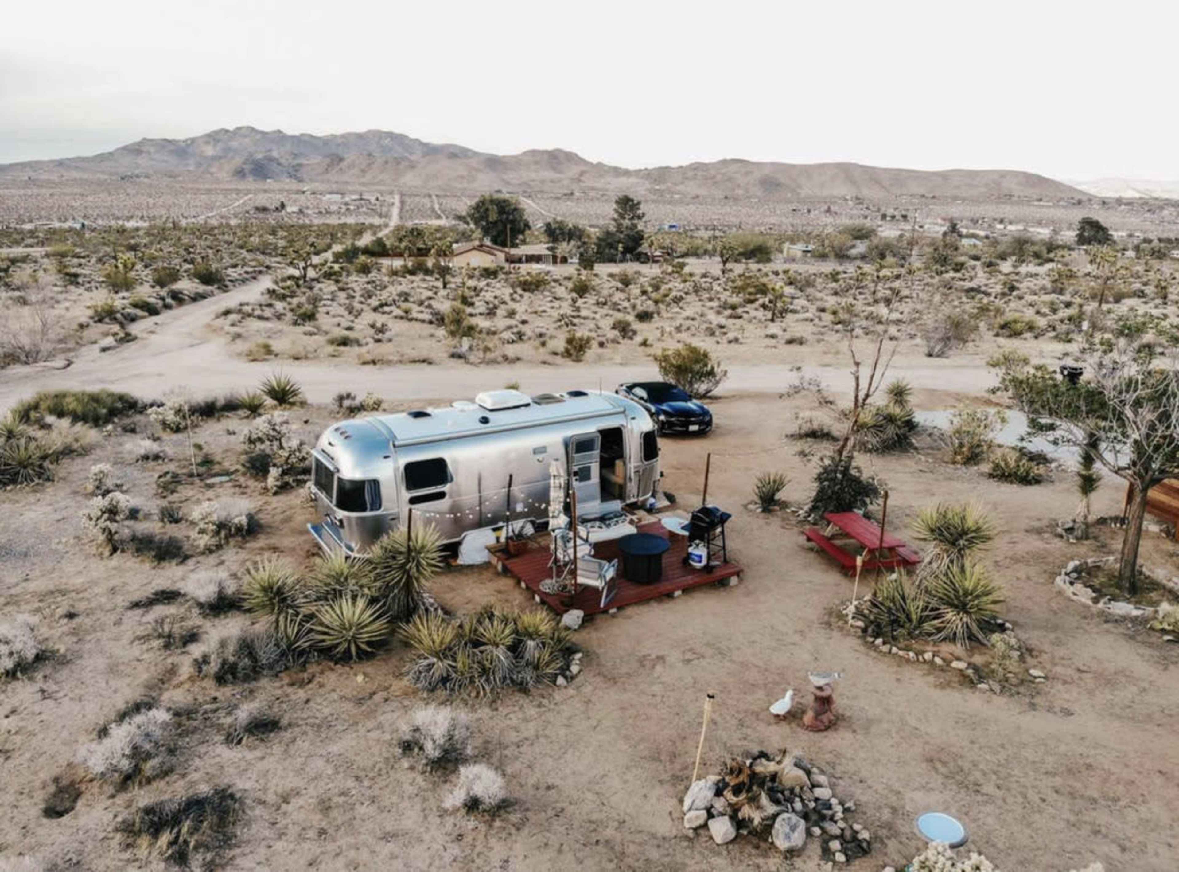 An Airstream trailer is parked on a wooden deck in a desert landscape surrounded by sparse vegetation and mountains in the background.