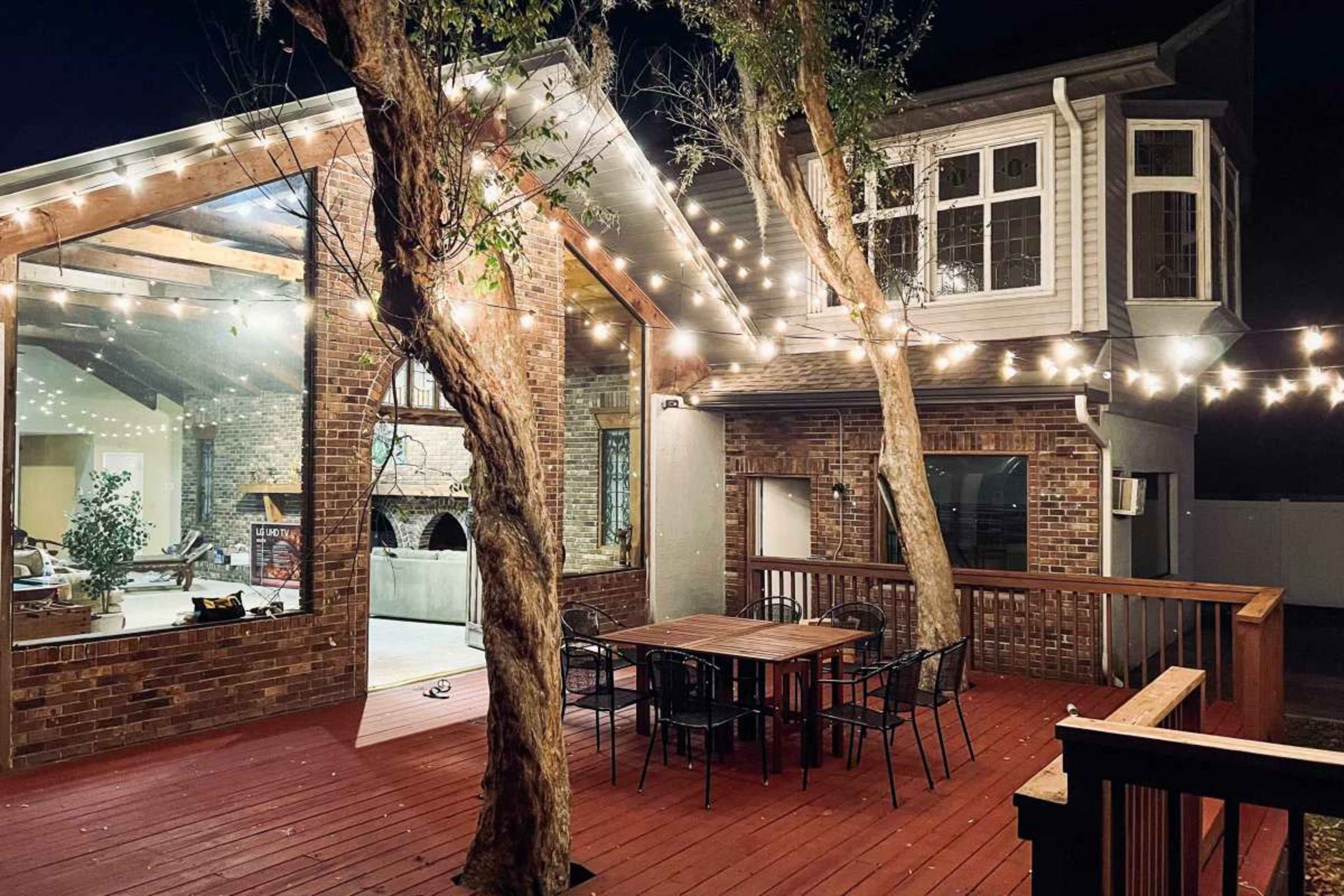 A wooden deck with a table and chairs is illuminated by string lights, adjacent to a brick house featuring large windows and visible interior spaces at night.