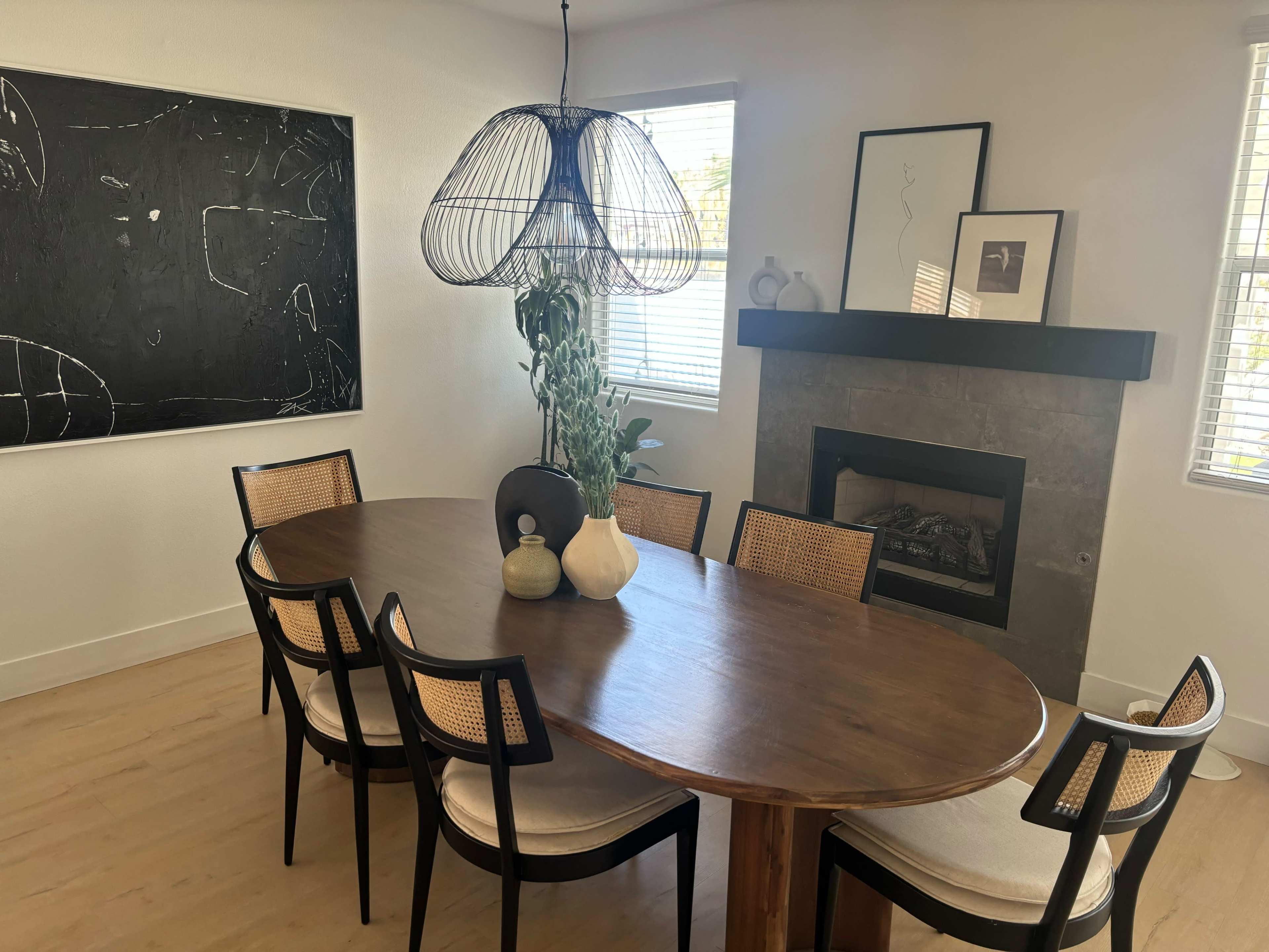 A modern dining room features a large oval wooden table surrounded by black and cane chairs, with a decorative pendant light and a stone fireplace in the background.
