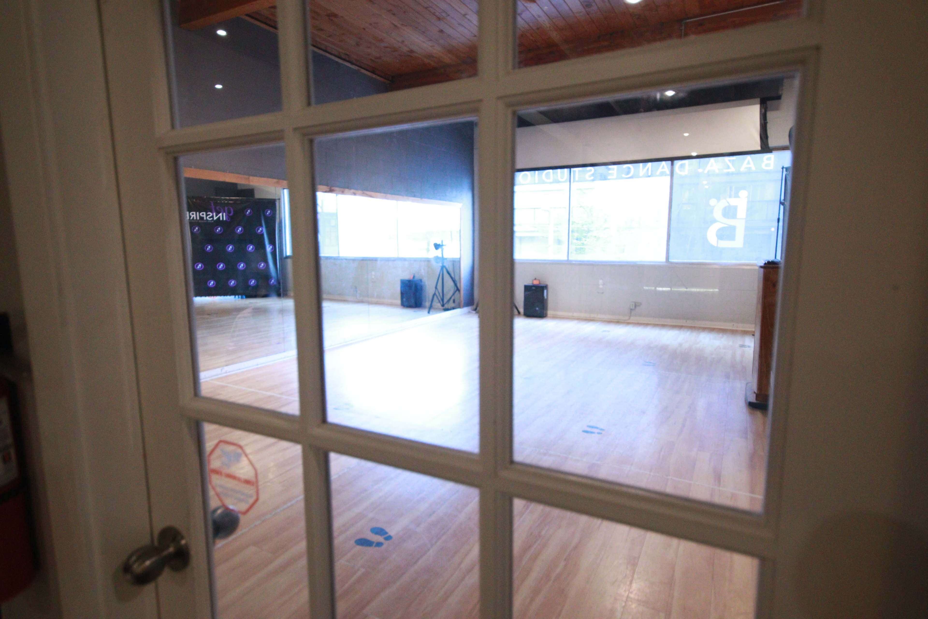 A dance studio viewed through a glass-paneled door, featuring a wooden floor and large windows illuminating the space.