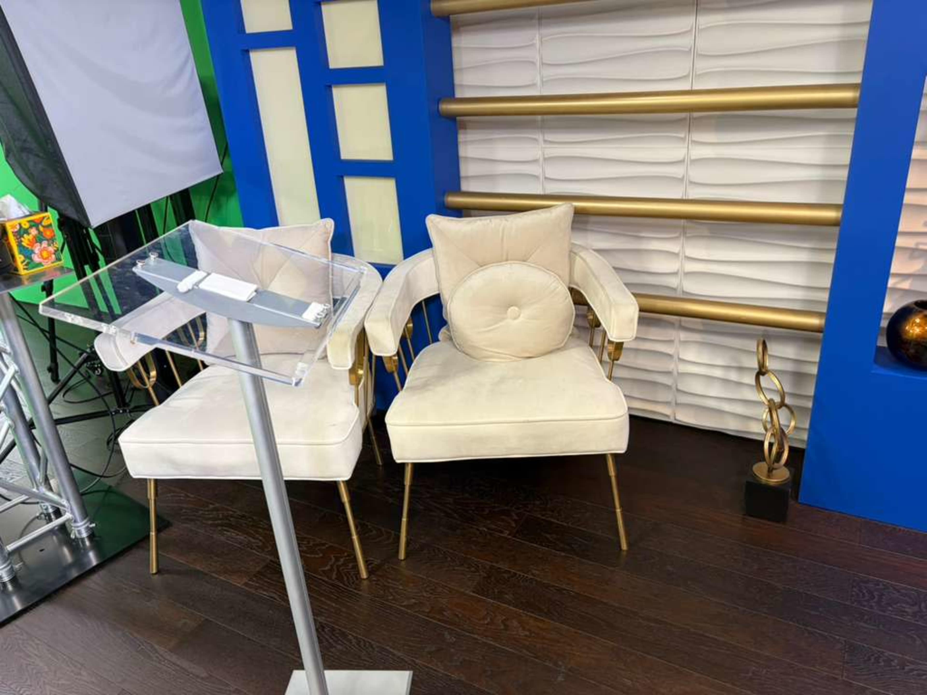 The image shows two cream-colored armchairs with gold accents positioned next to a clear acrylic podium, set against a backdrop of blue and textured white panels.