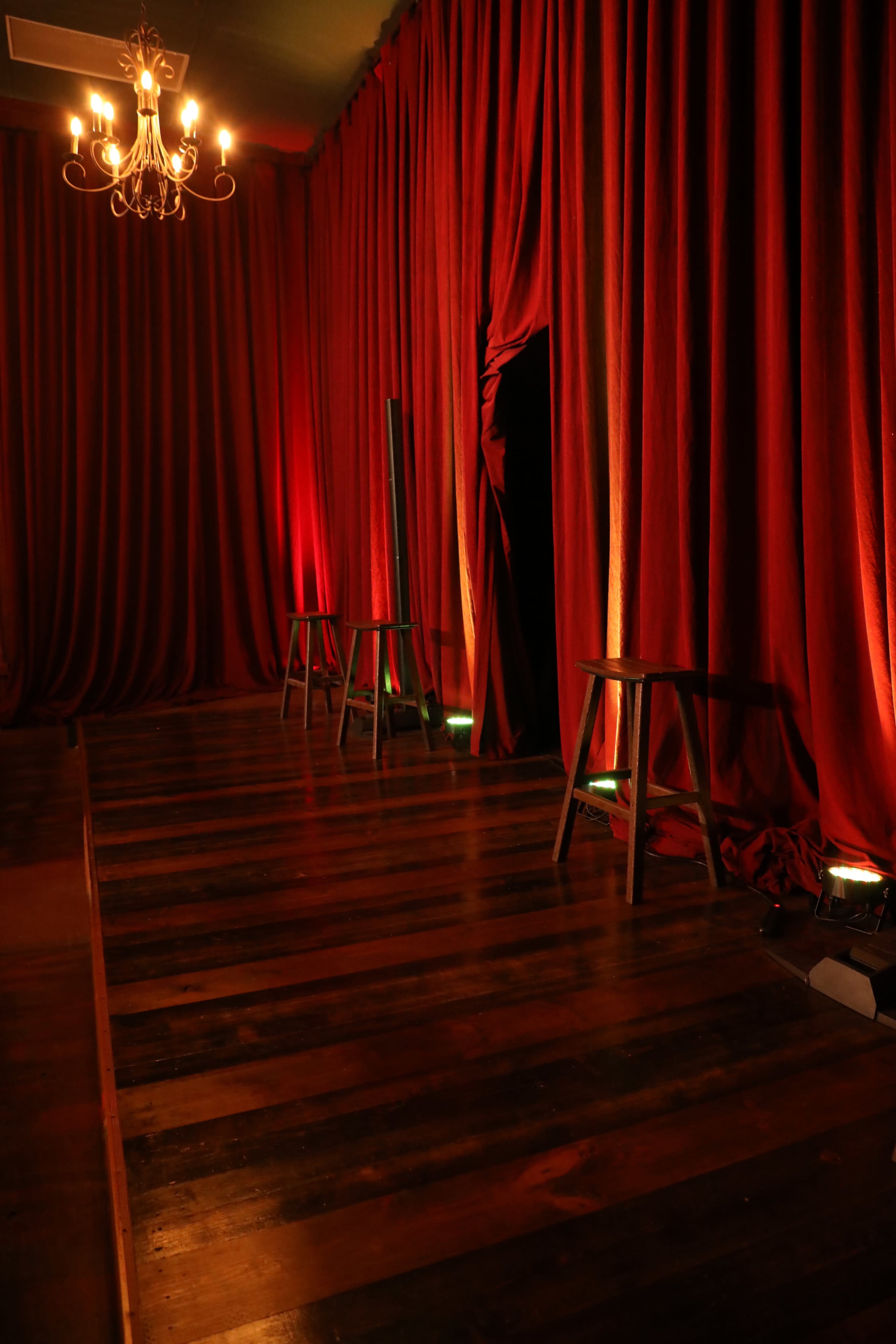 The image depicts a dimly lit stage with red velvet curtains, wooden flooring, and two wooden stools positioned in front.