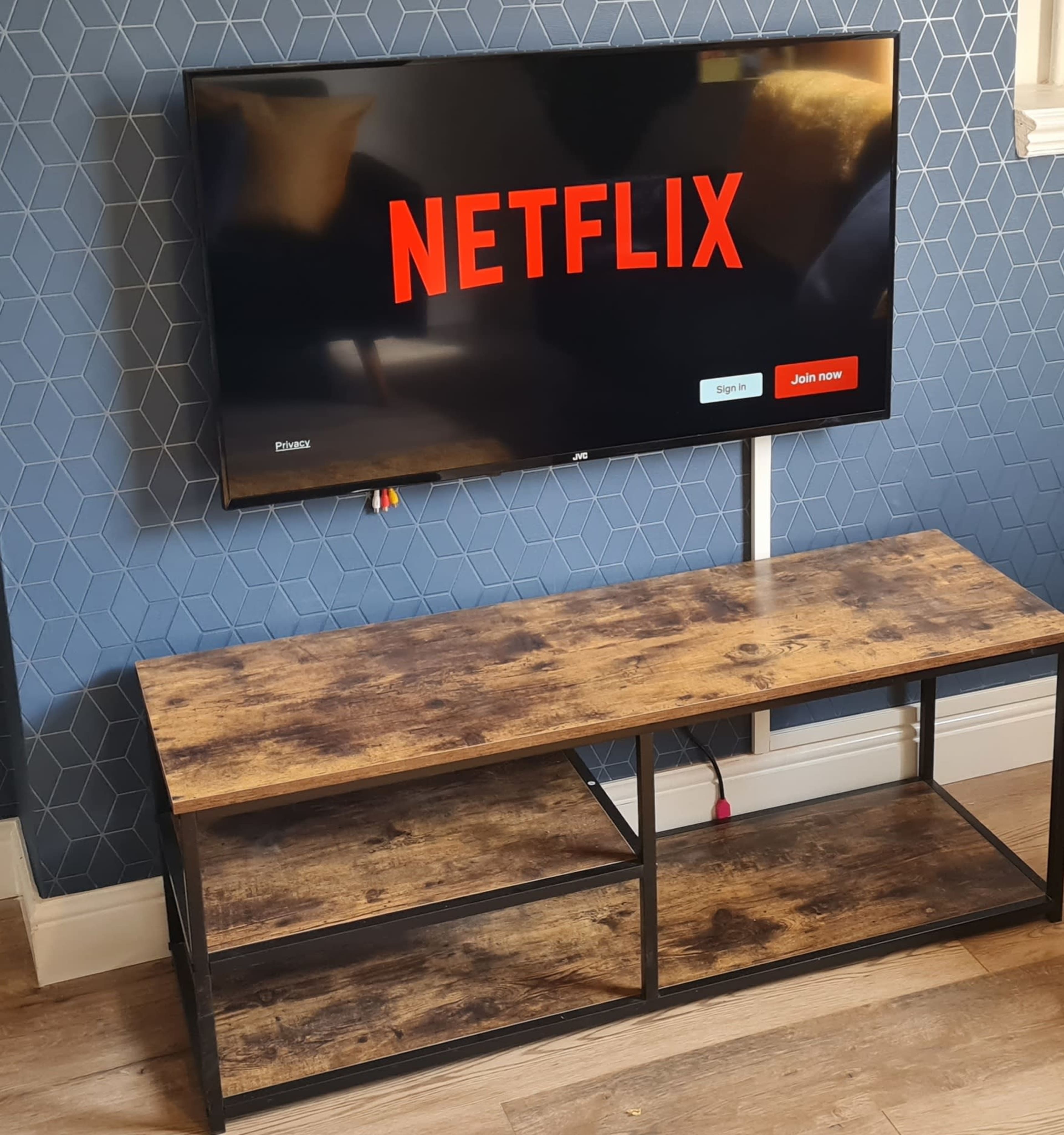 A flat-screen TV displaying the Netflix logo is mounted on a wall above a wooden stand with two open shelves.