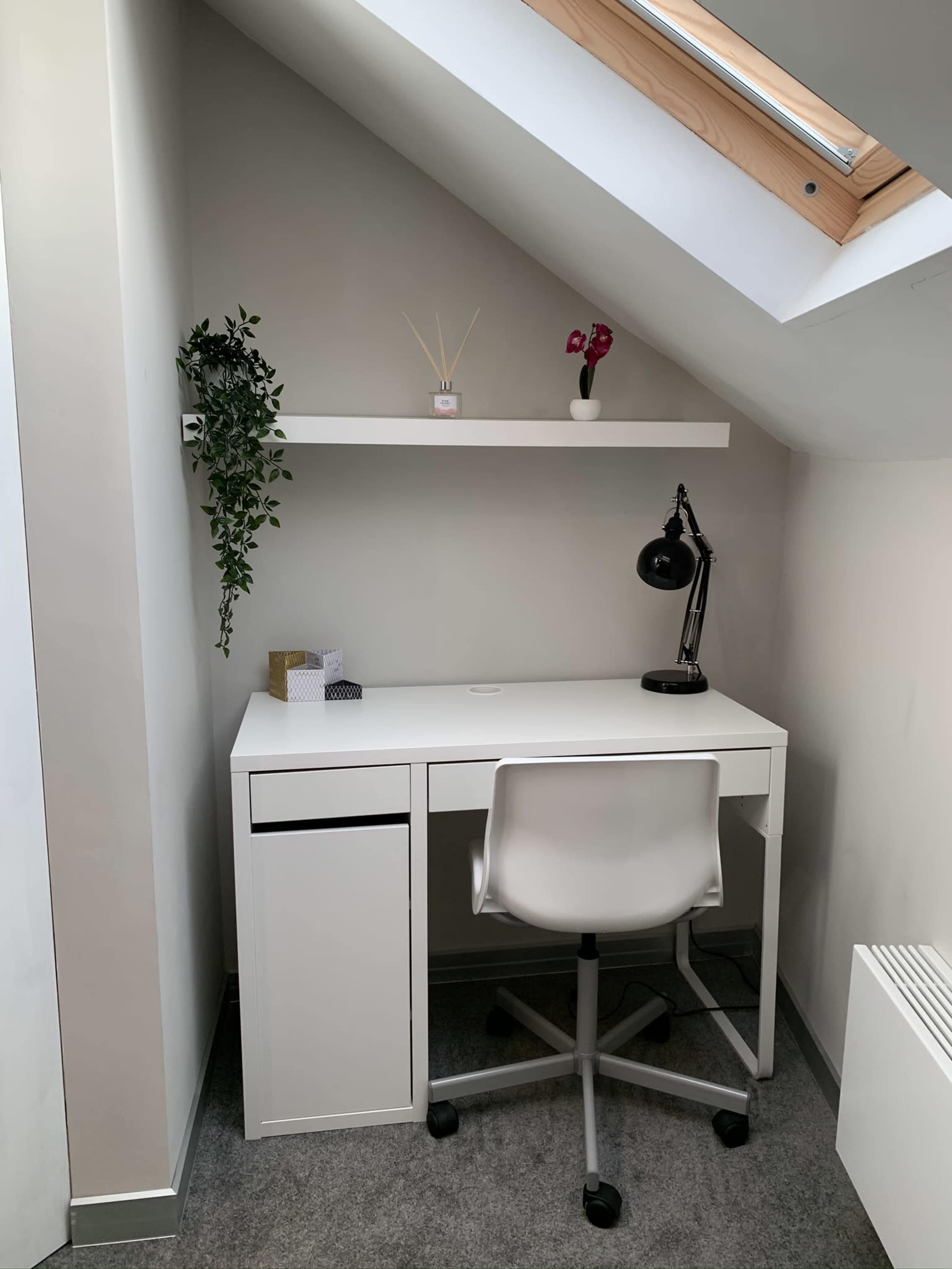 A minimalist desk setup in a corner, featuring a white desk, a swivel chair, a lamp, and decorative items on a shelf above.