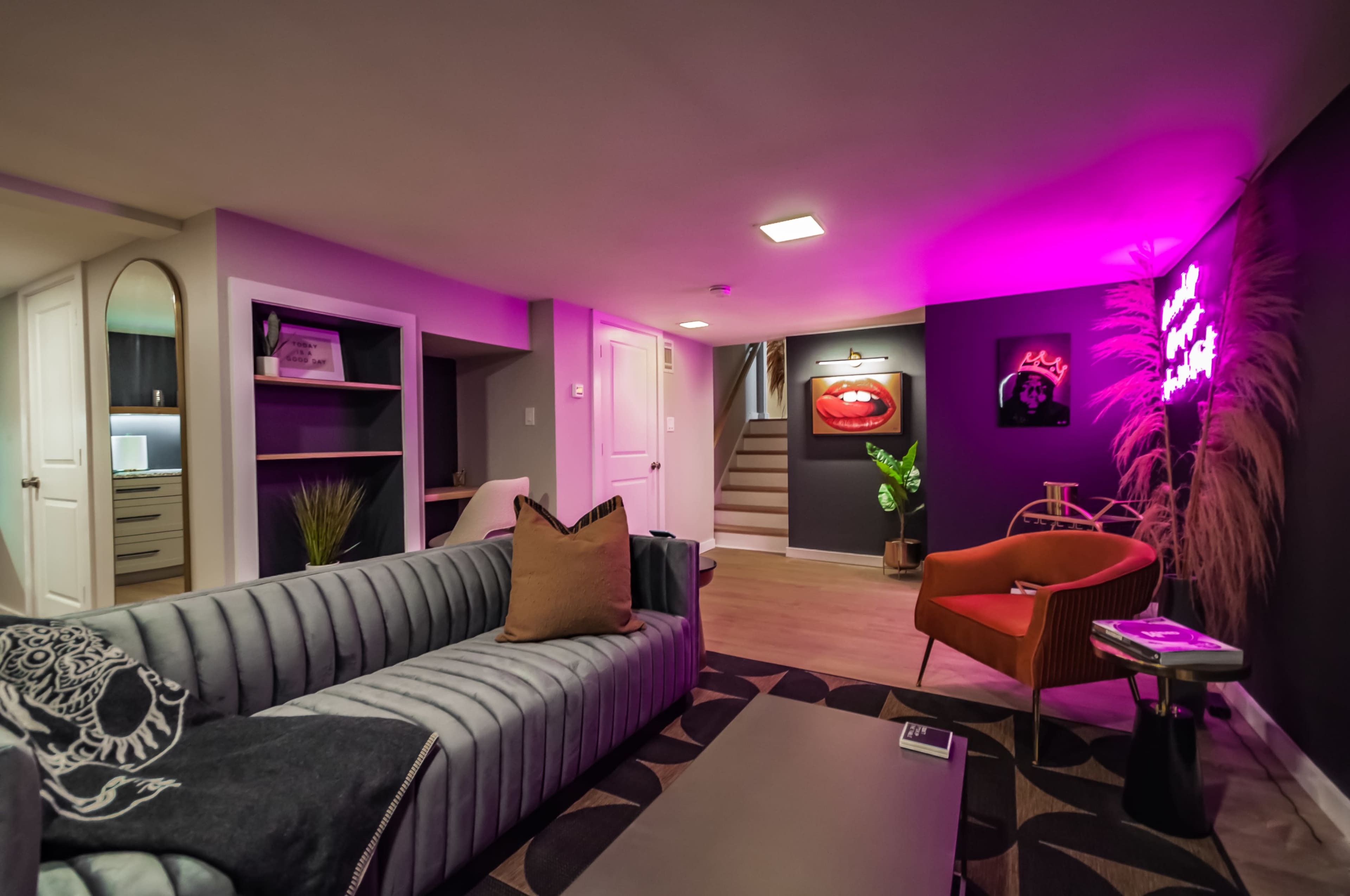 A modern basement lounge area with a gray sofa, a red armchair, pink ambient lighting, and decorative art pieces on the walls.