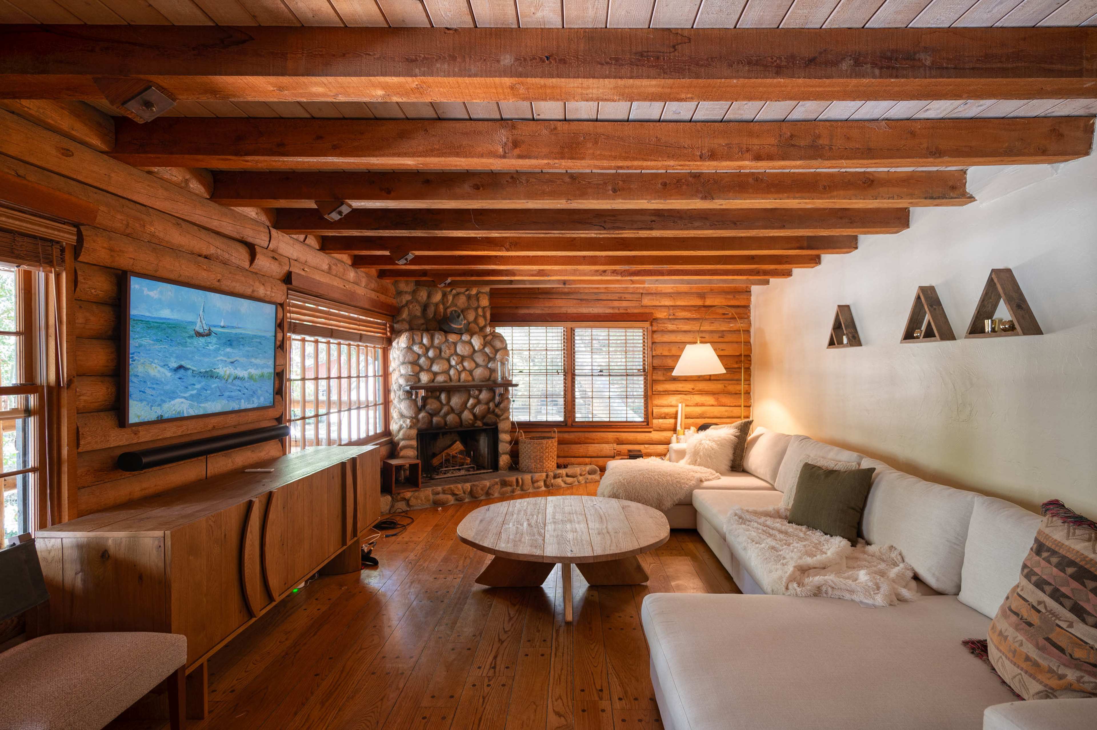 The interior of a rustic living room features wood-paneled walls, a stone fireplace, a large sectional sofa, and a circular coffee table.