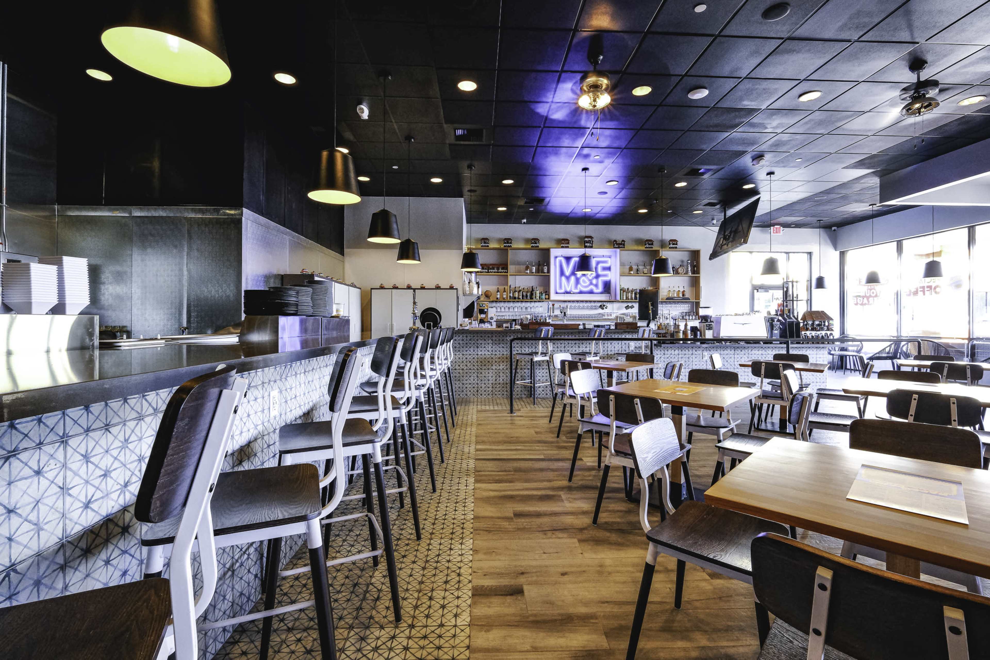 The image shows a modern restaurant interior featuring a bar area, wooden tables, and high-backed chairs with a patterned tile wall and shelves stocked with liquor.