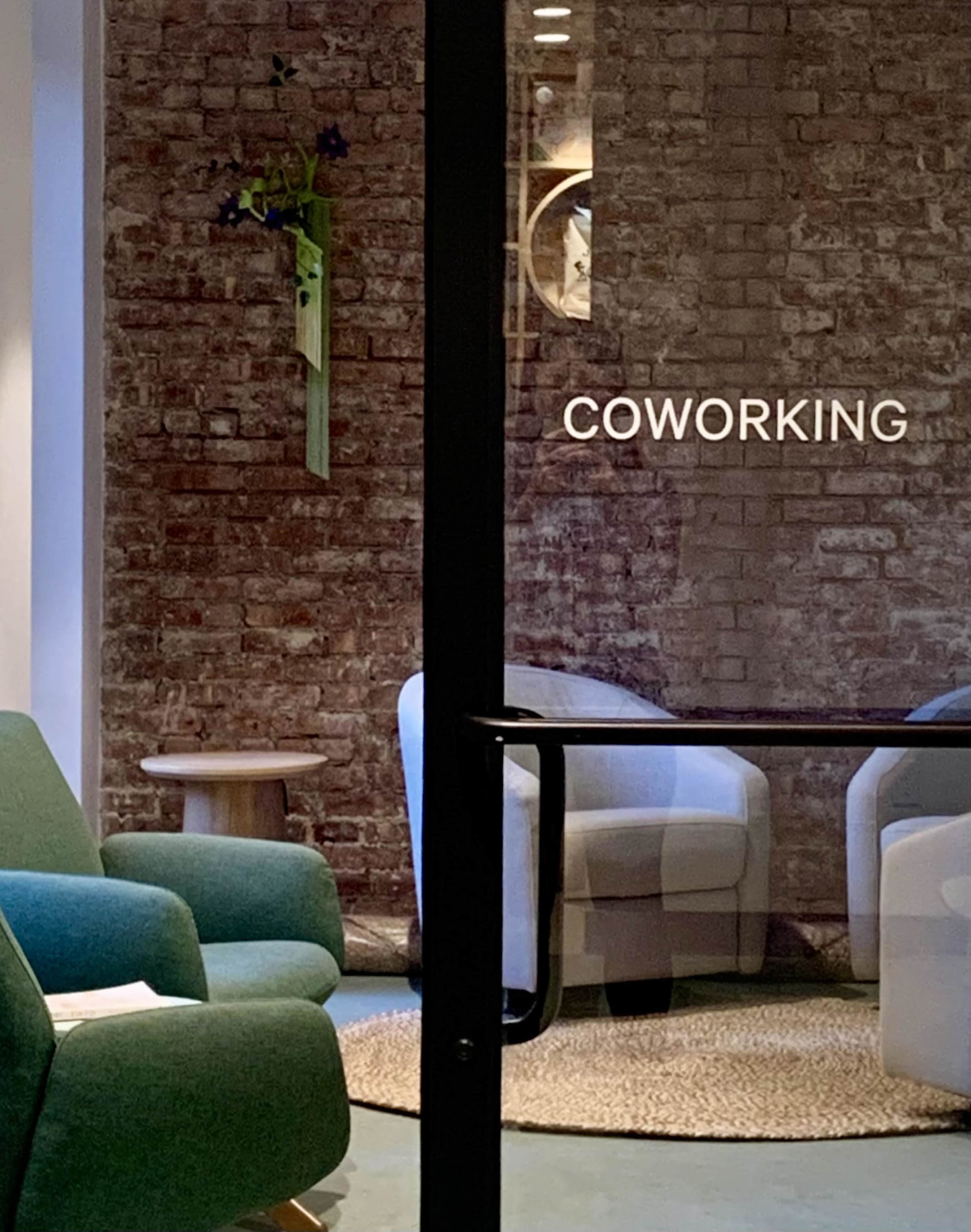 The image shows a coworking space featuring green upholstered chairs, a round table, and a textured brick wall.