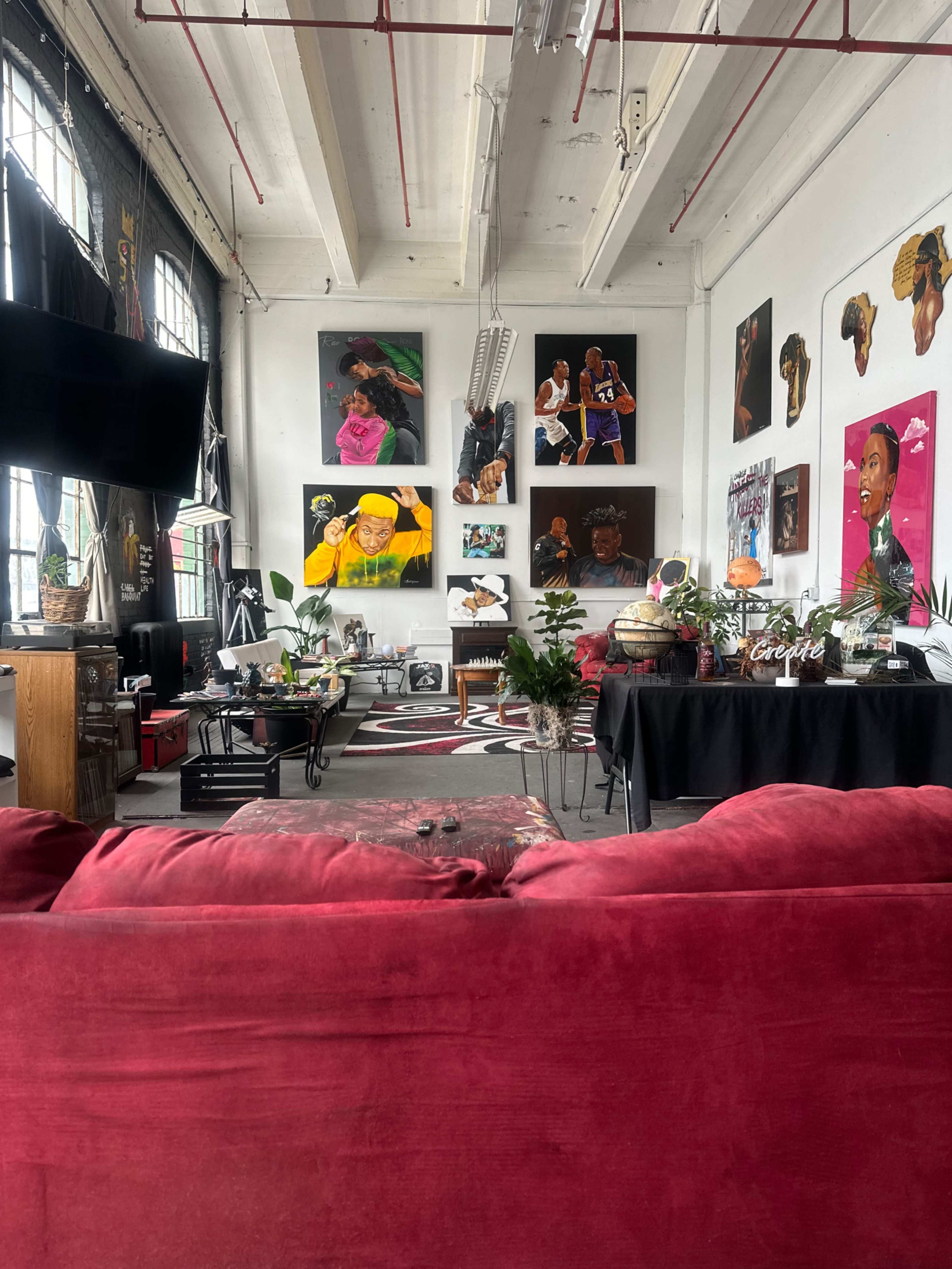 The image shows a lively art studio featuring bright paintings of various figures on the walls, with a red couch in the foreground and art supplies scattered around.