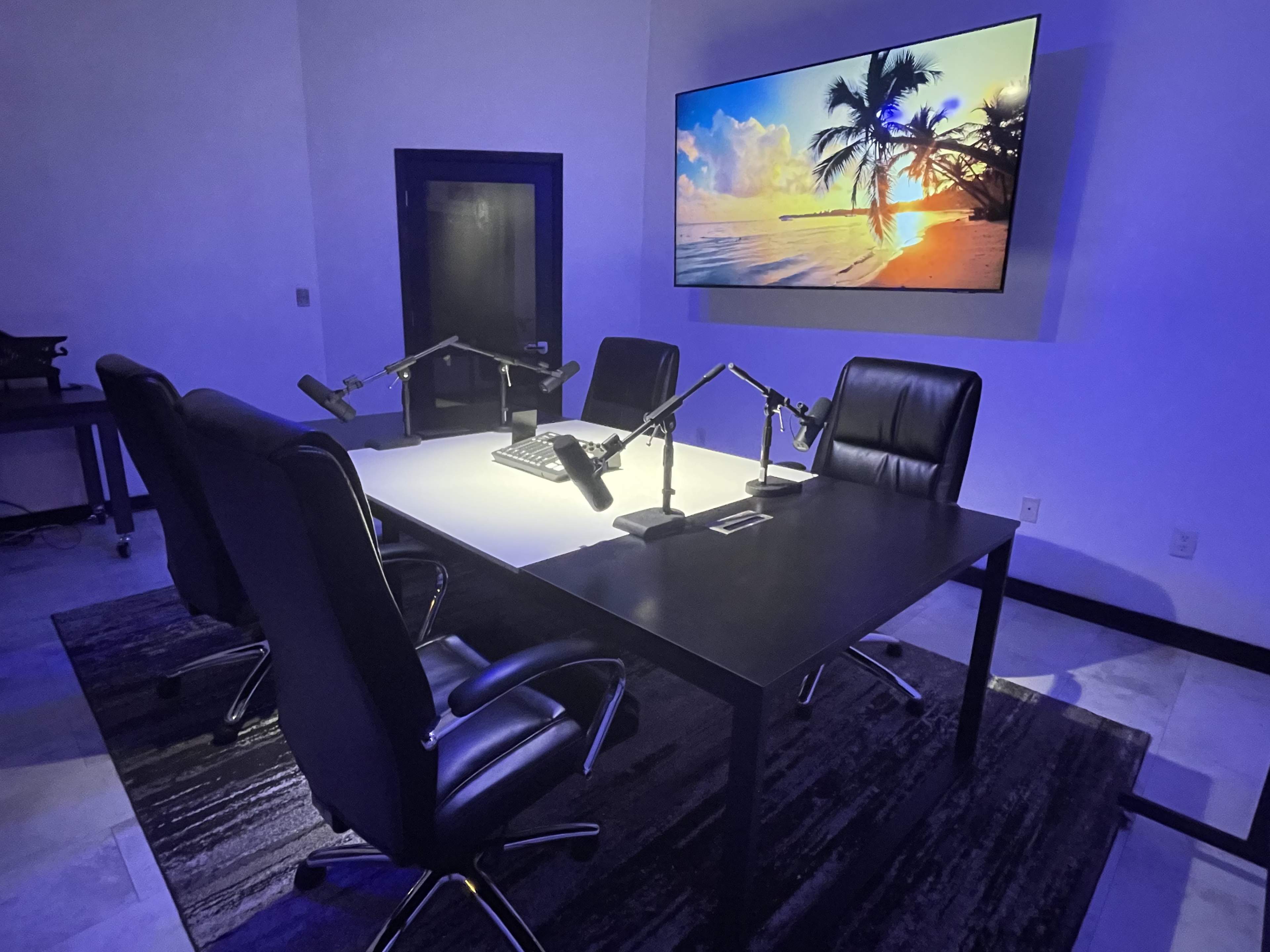 A modern conference room features a large table with four black chairs, illuminated by overhead light, and a large screen displaying a beach scene.