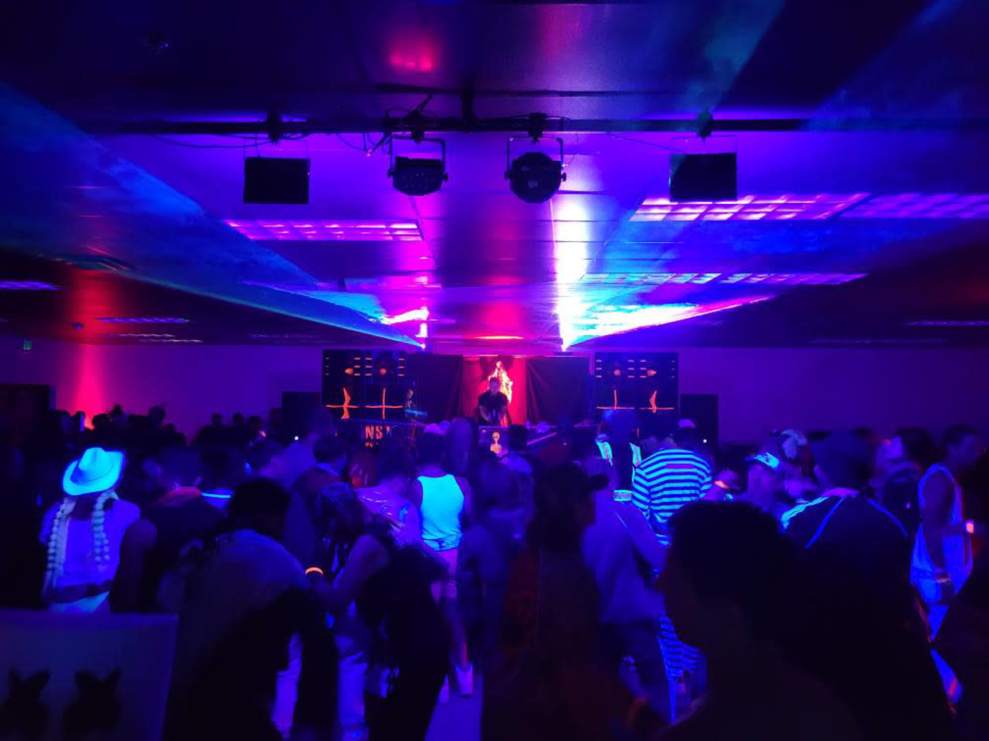A crowded dance floor is illuminated by colorful lights, with a performer on stage in the background.