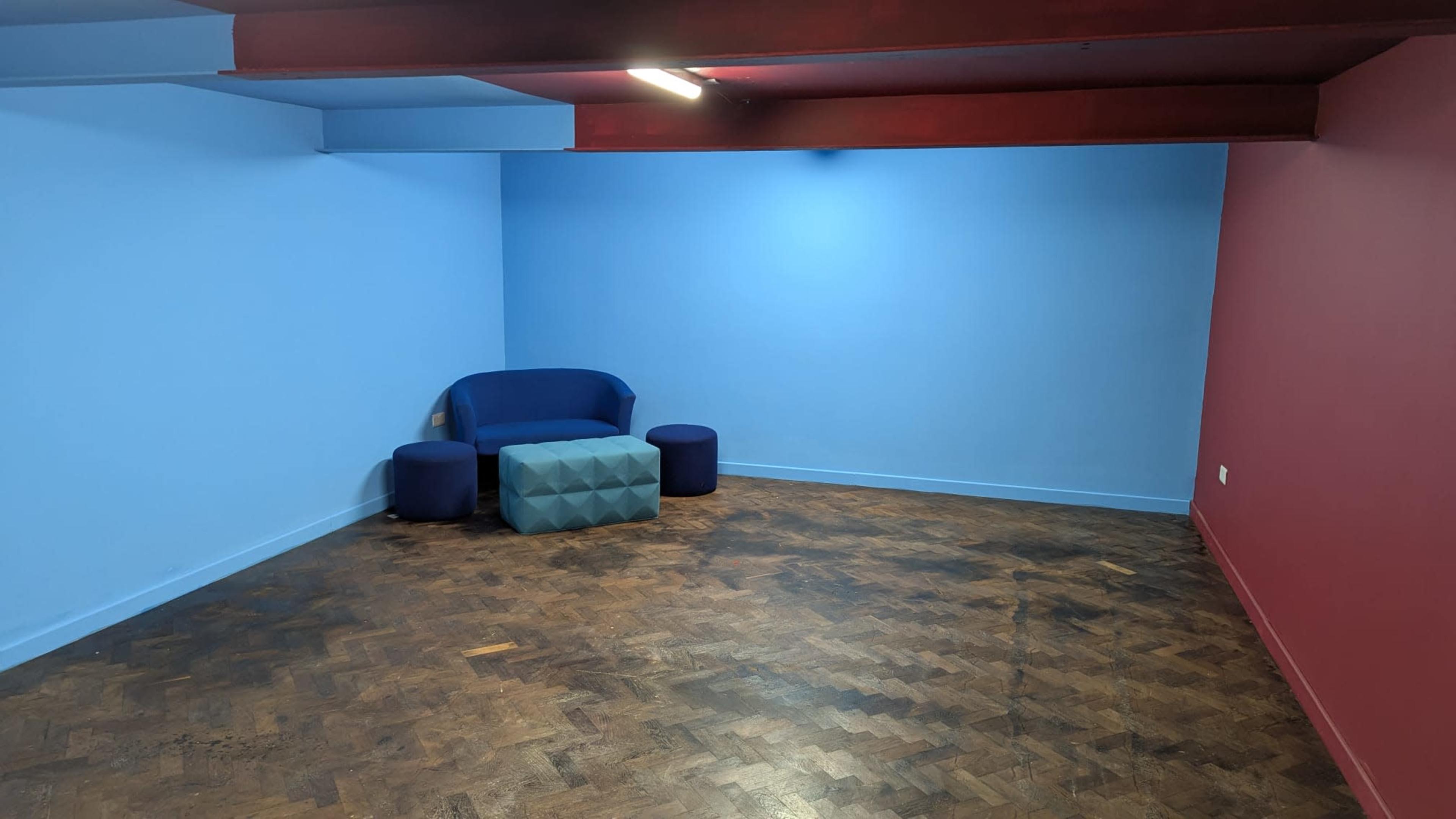 The image shows a sparsely furnished room with blue and red walls, featuring a blue couch, a geometric coffee table, and two round stools on a wooden floor.