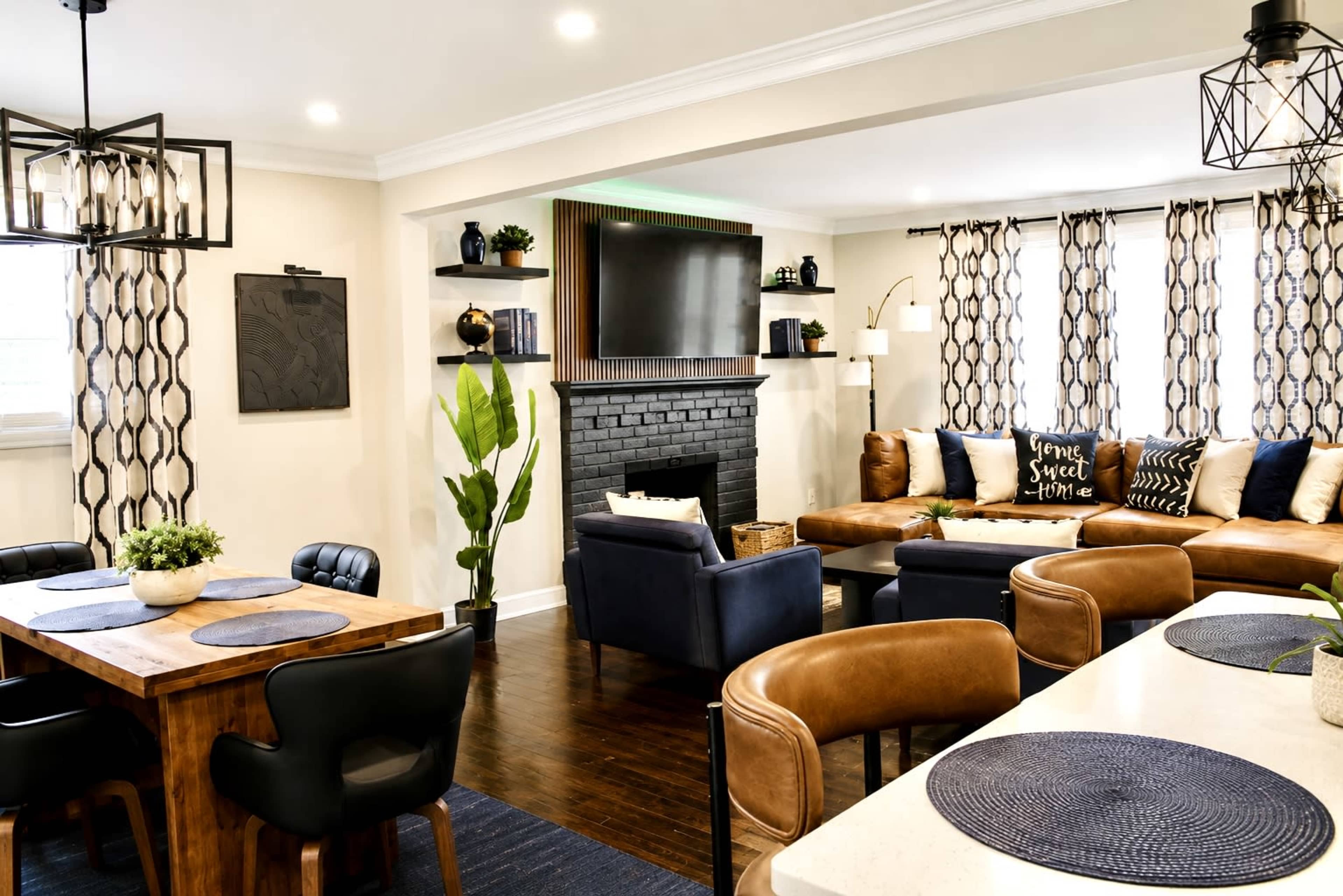 The image shows a modern living room and dining area featuring a sectional sofa, a large television mounted above a brick fireplace, and a wooden dining table with black and brown chairs.