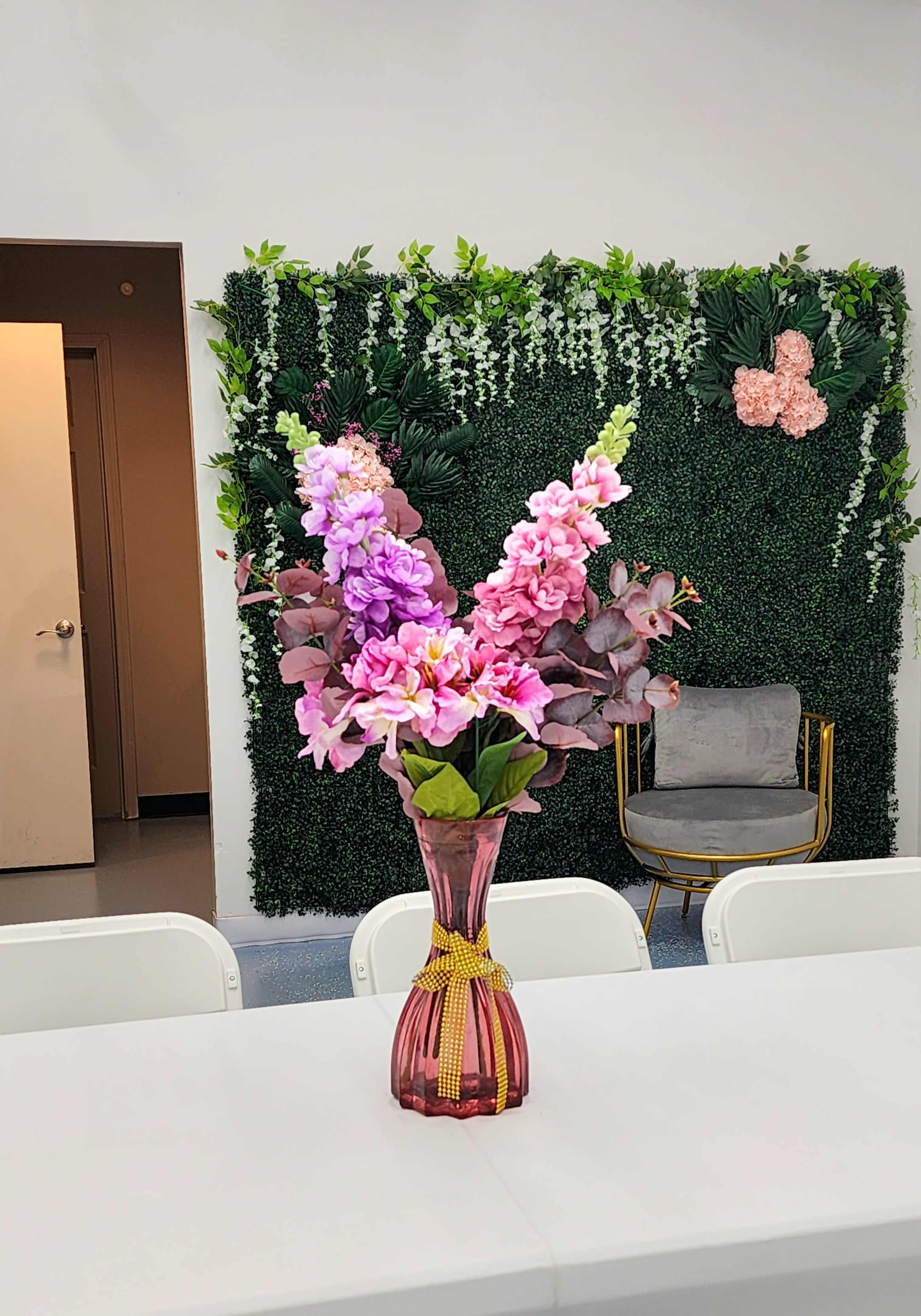 A vase of pink and purple flowers sits on a white table in front of a green wall adorned with plants and decorative accents.