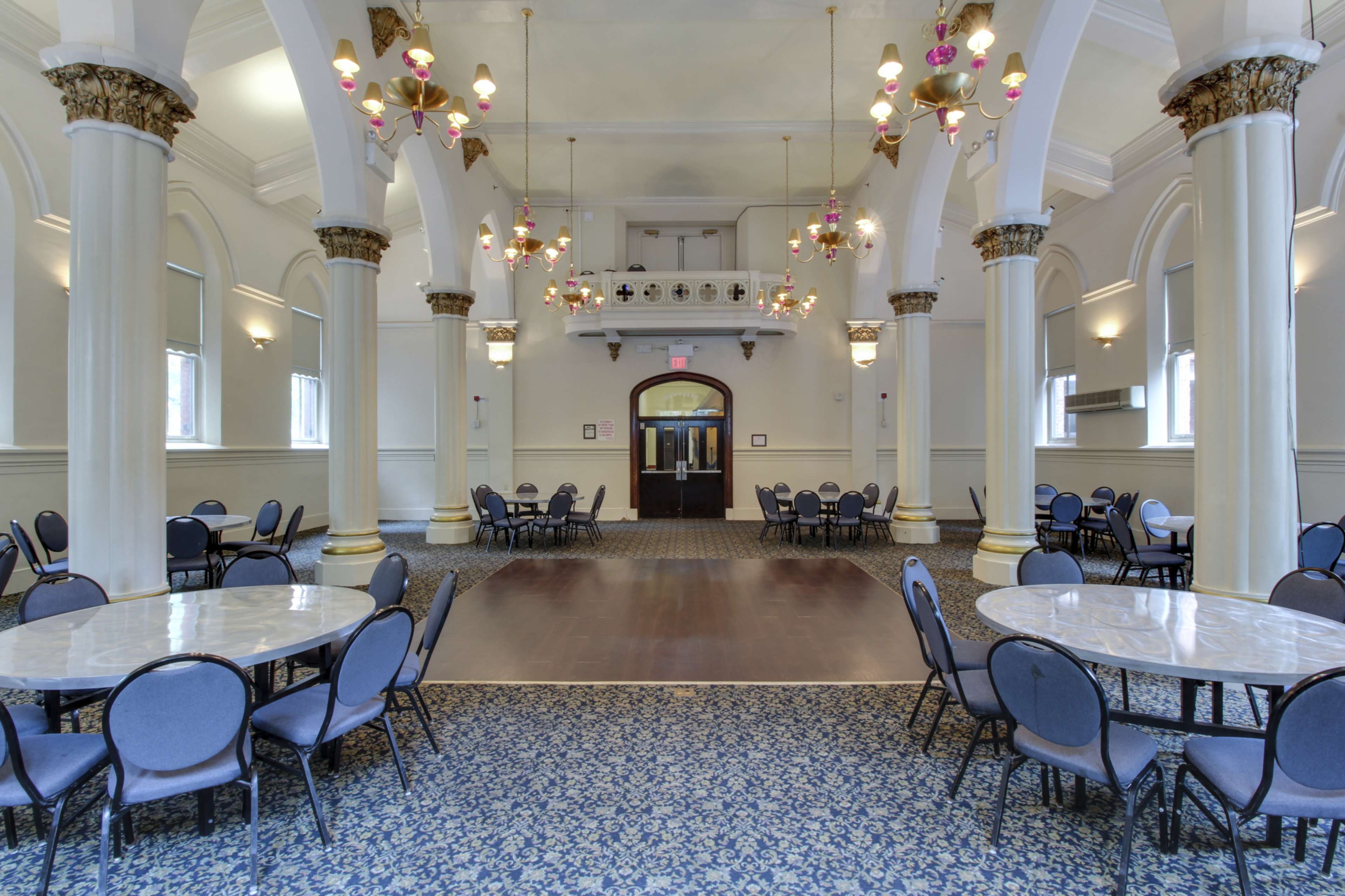 The image shows a large, elegant hall with round tables and chairs arranged across a patterned carpet, featuring high ceilings and decorative chandeliers.