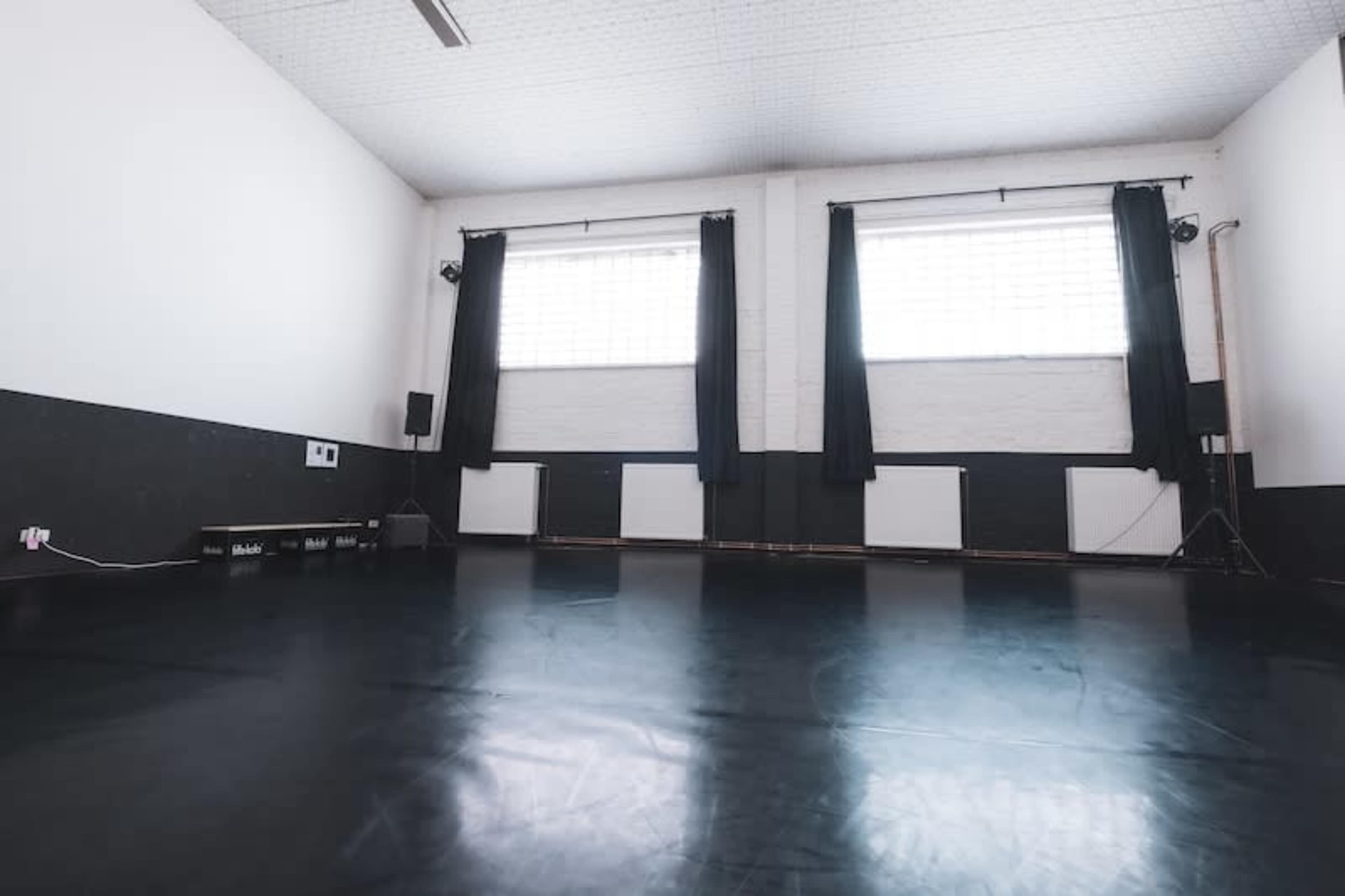 An empty dance studio with black flooring, large windows covered by dark curtains, and minimal furnishings.