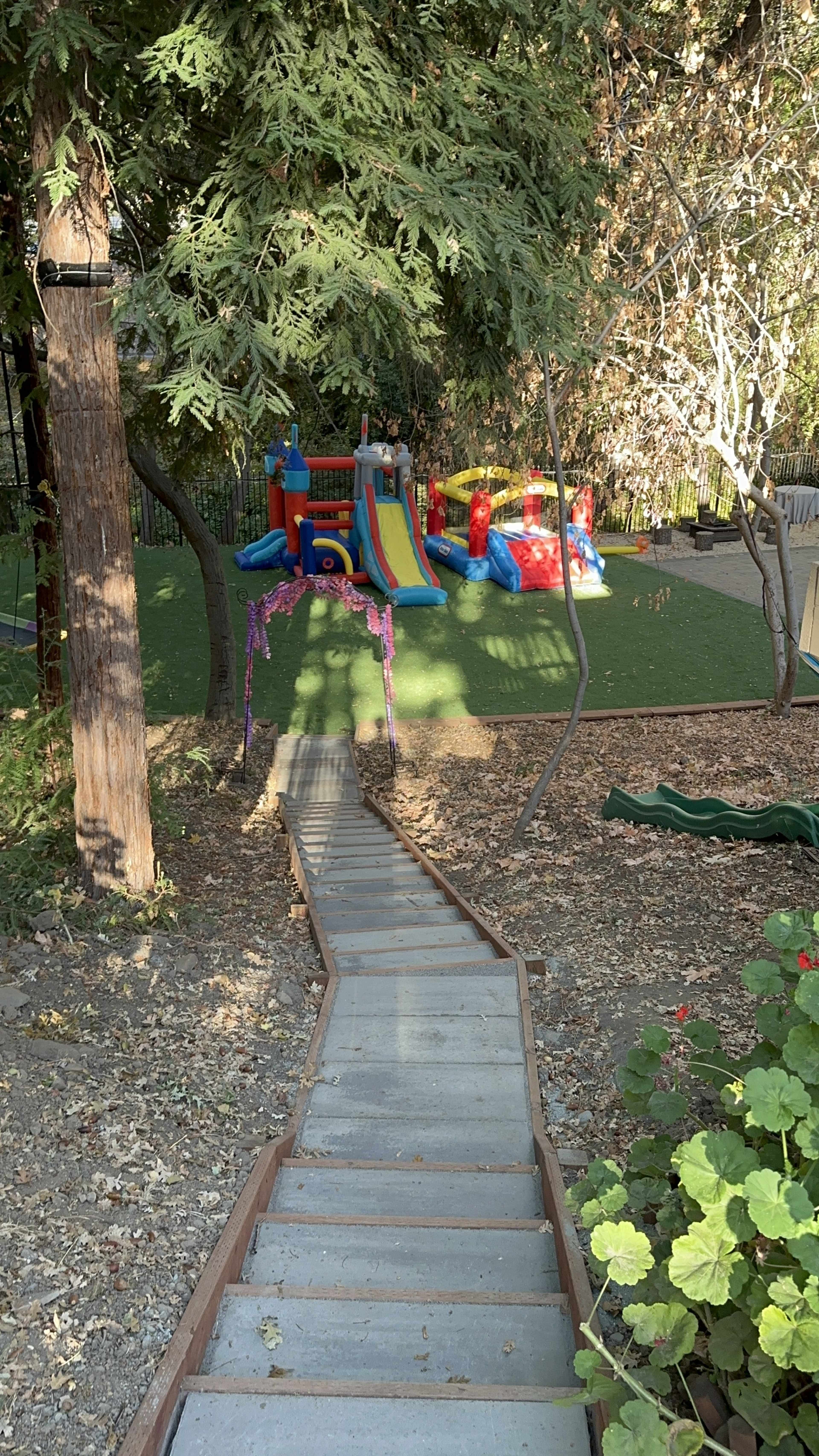A wooden staircase leads down to a colorful playground featuring inflatable slides and play structures surrounded by trees.