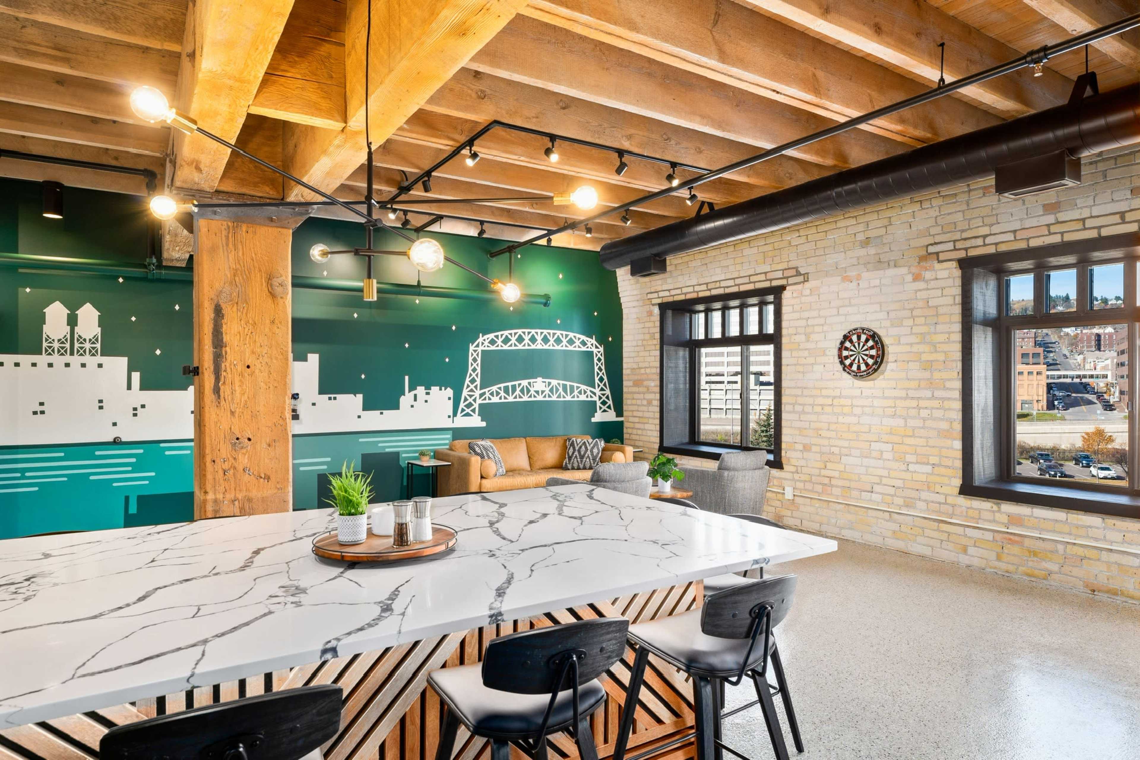 A modern loft-style living space featuring a marble countertop island, exposed wooden beams, a green cityscape mural, and a dartboard on the wall.