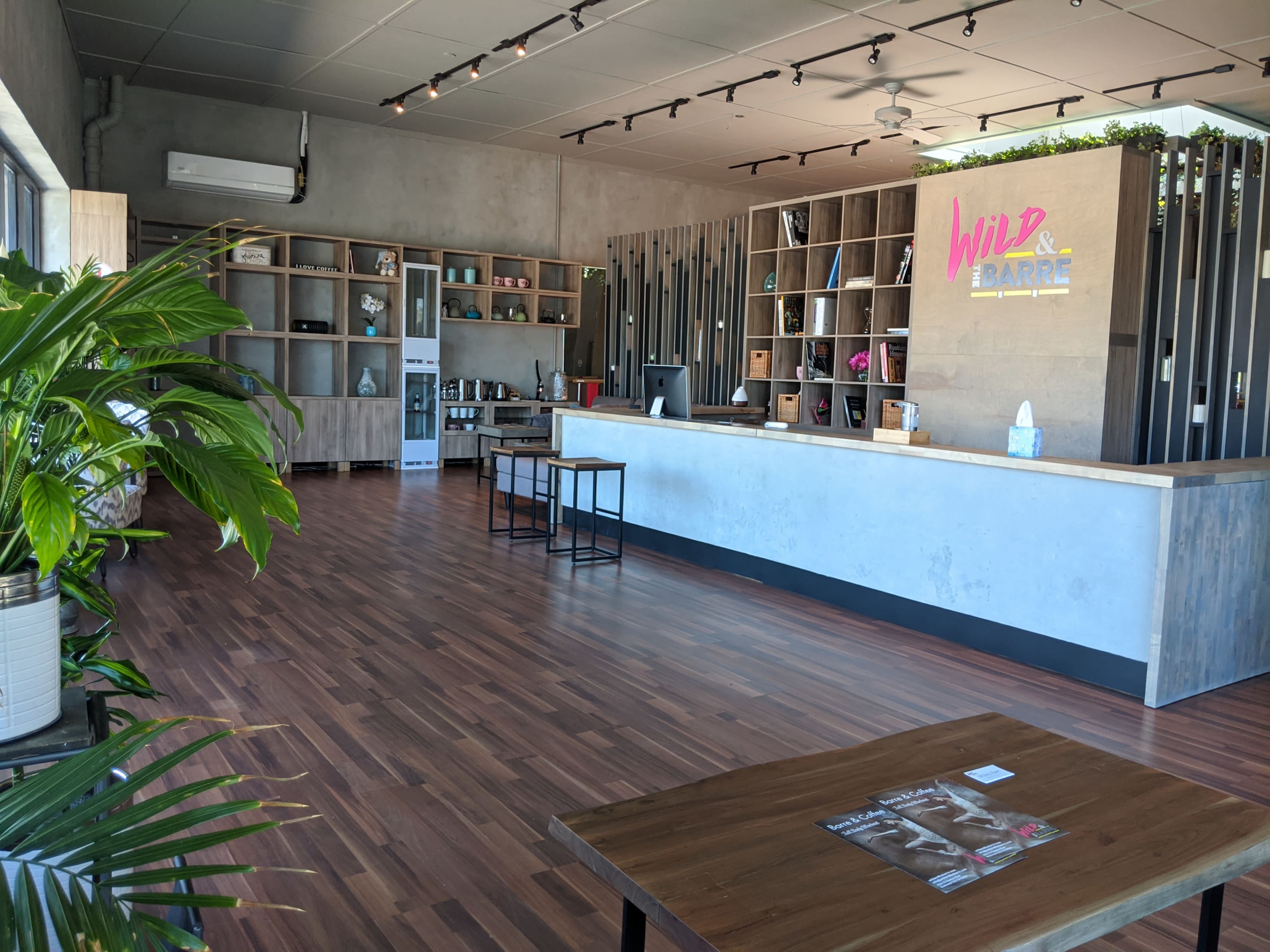 A modern, spacious interior of a café or bar with wooden flooring, a minimalistic counter, shelves displaying various items, and plants.