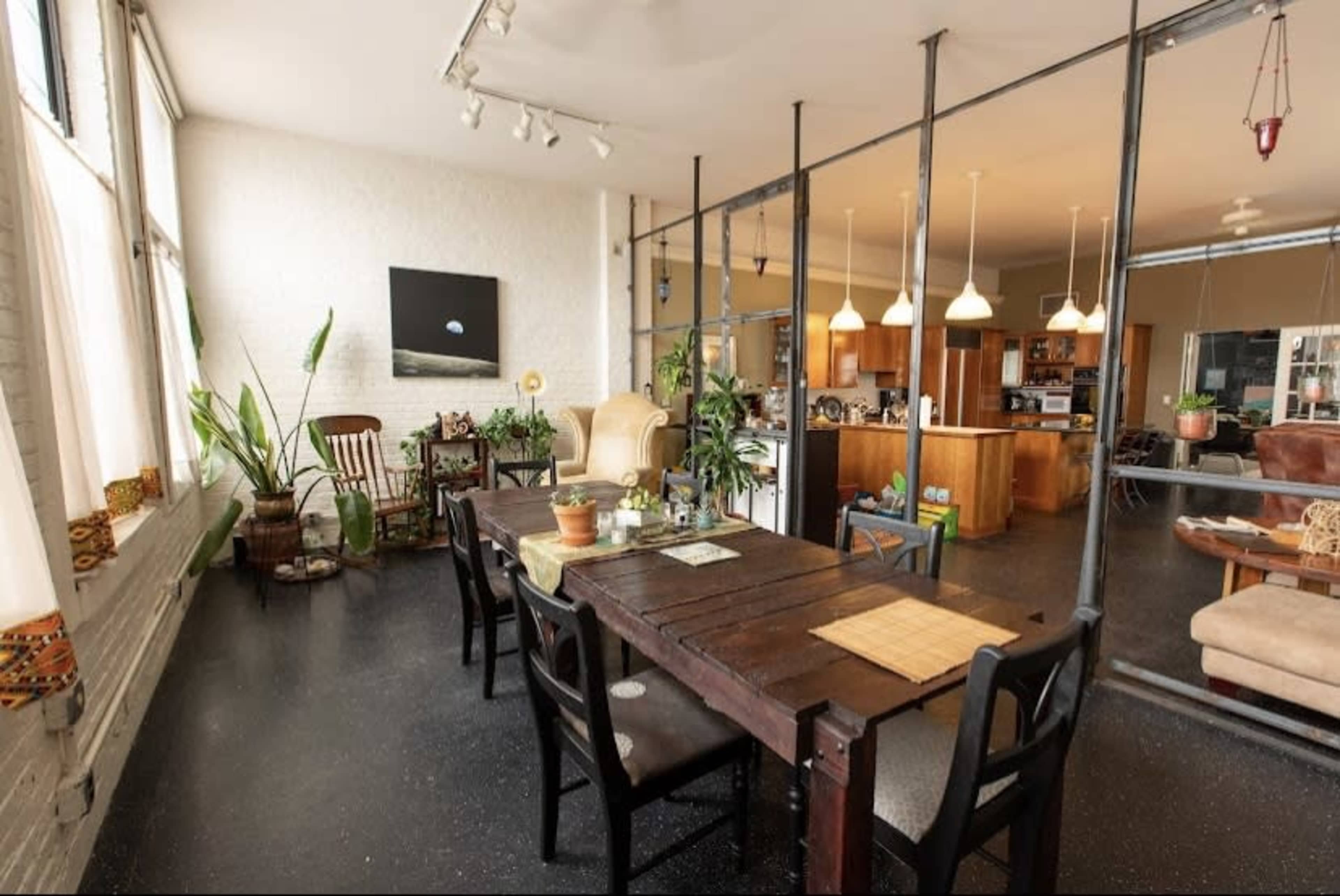 An open-plan living space with a wooden dining table surrounded by black chairs, a kitchenette visible in the background, and large windows allowing natural light to enter.