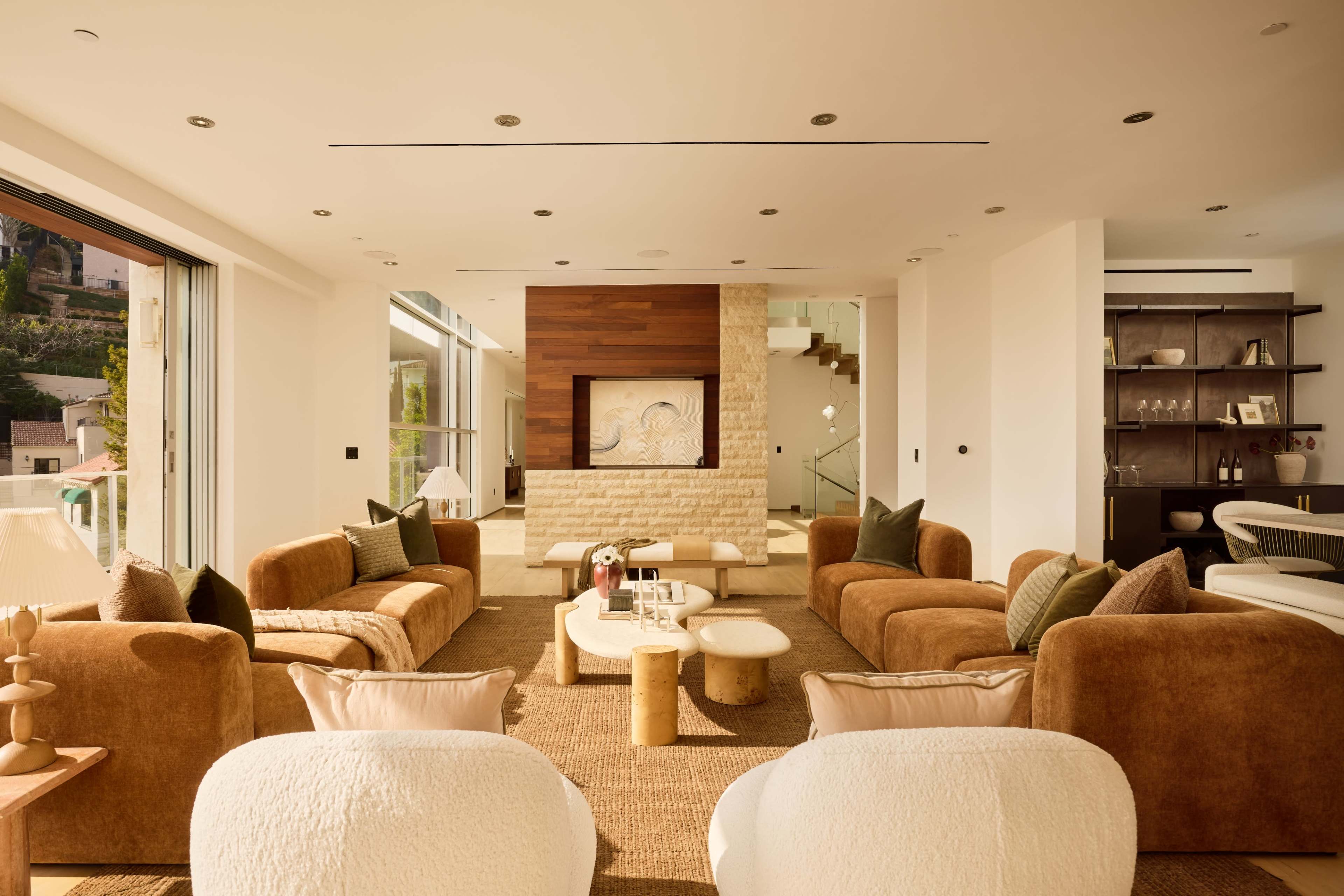 The image shows a modern living room featuring multiple brown sofas, a light-colored stone fireplace, and large windows that allow natural light to fill the space.