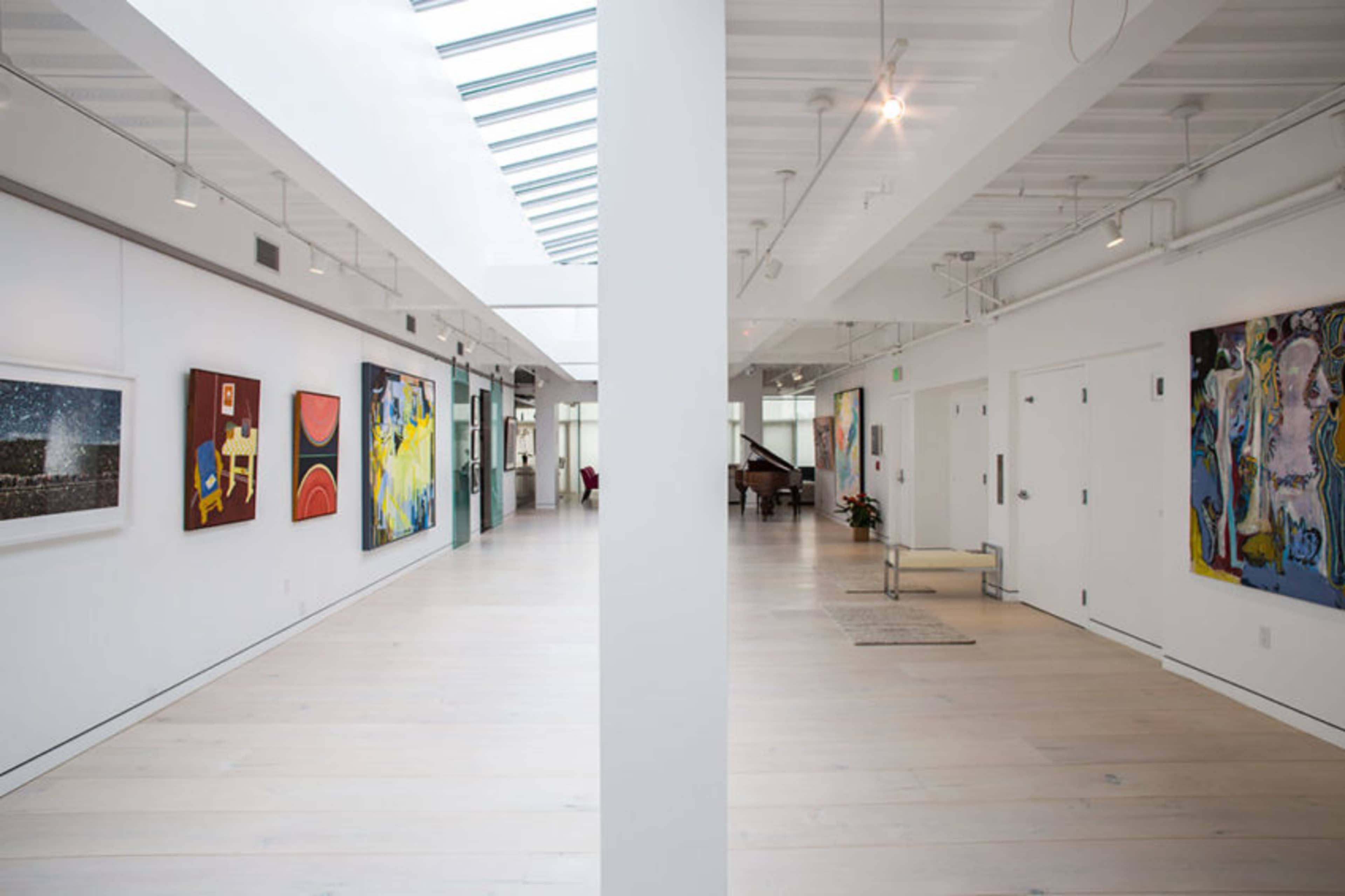 A spacious, brightly lit gallery hallway featuring various colorful artworks displayed on white walls, with a grand piano visible in the background.