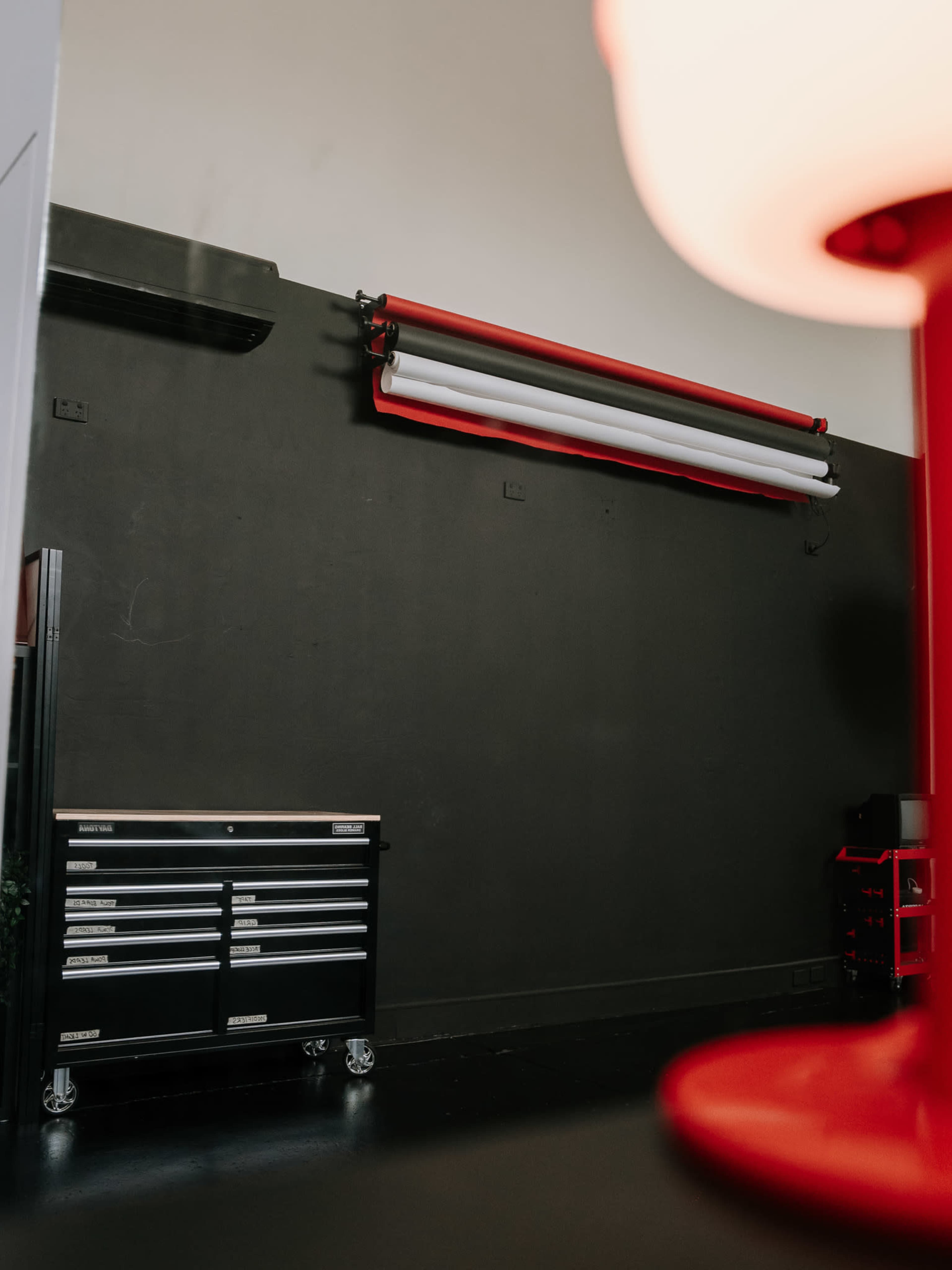 The image shows a workspace with a black wall, a mounted roll of paper backdrop, a metal tool chest on wheels, and a red lamp in the foreground.