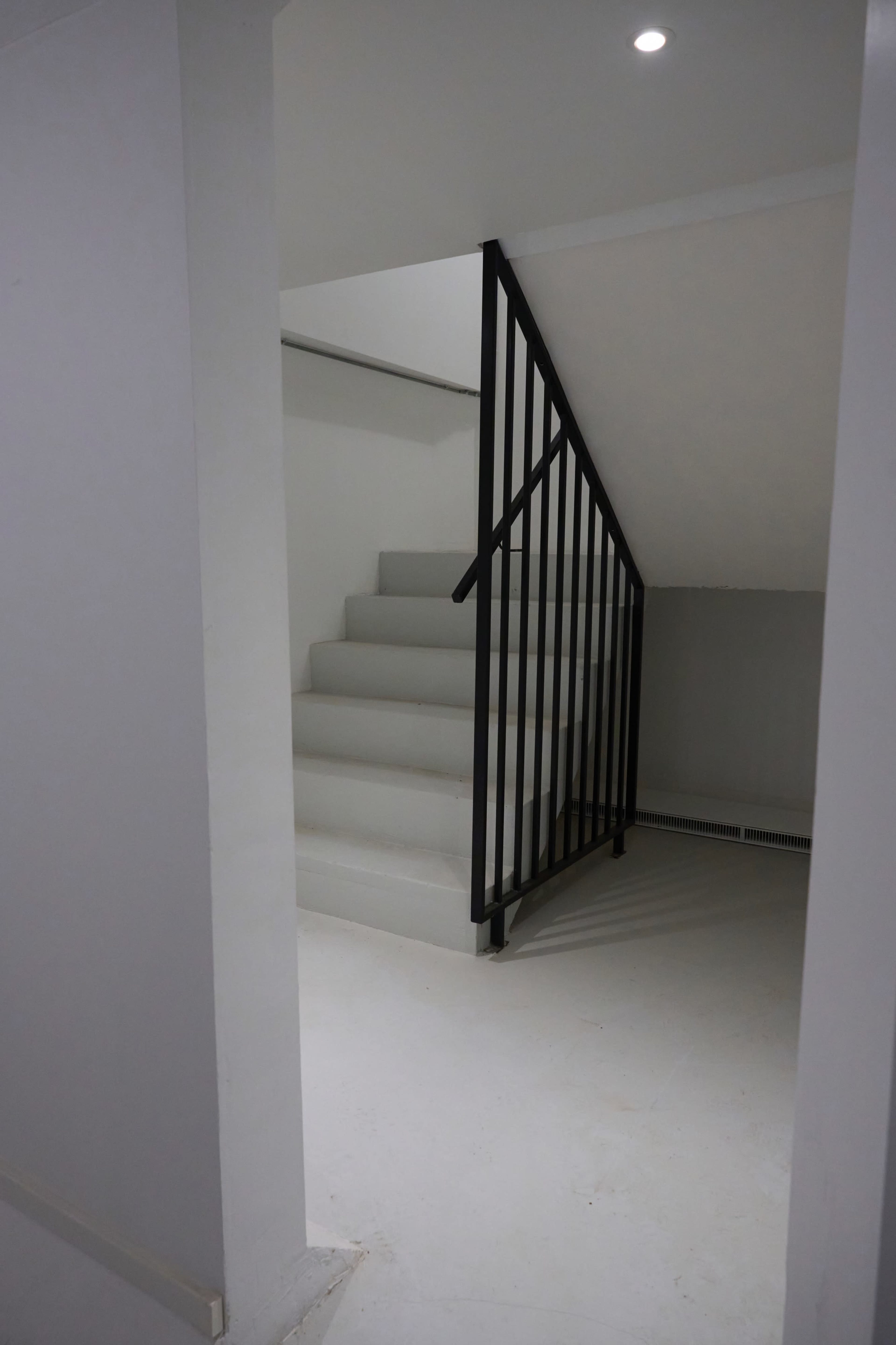 A staircase with a black railing leads to an upper level in a minimally designed space with light-colored walls and flooring.