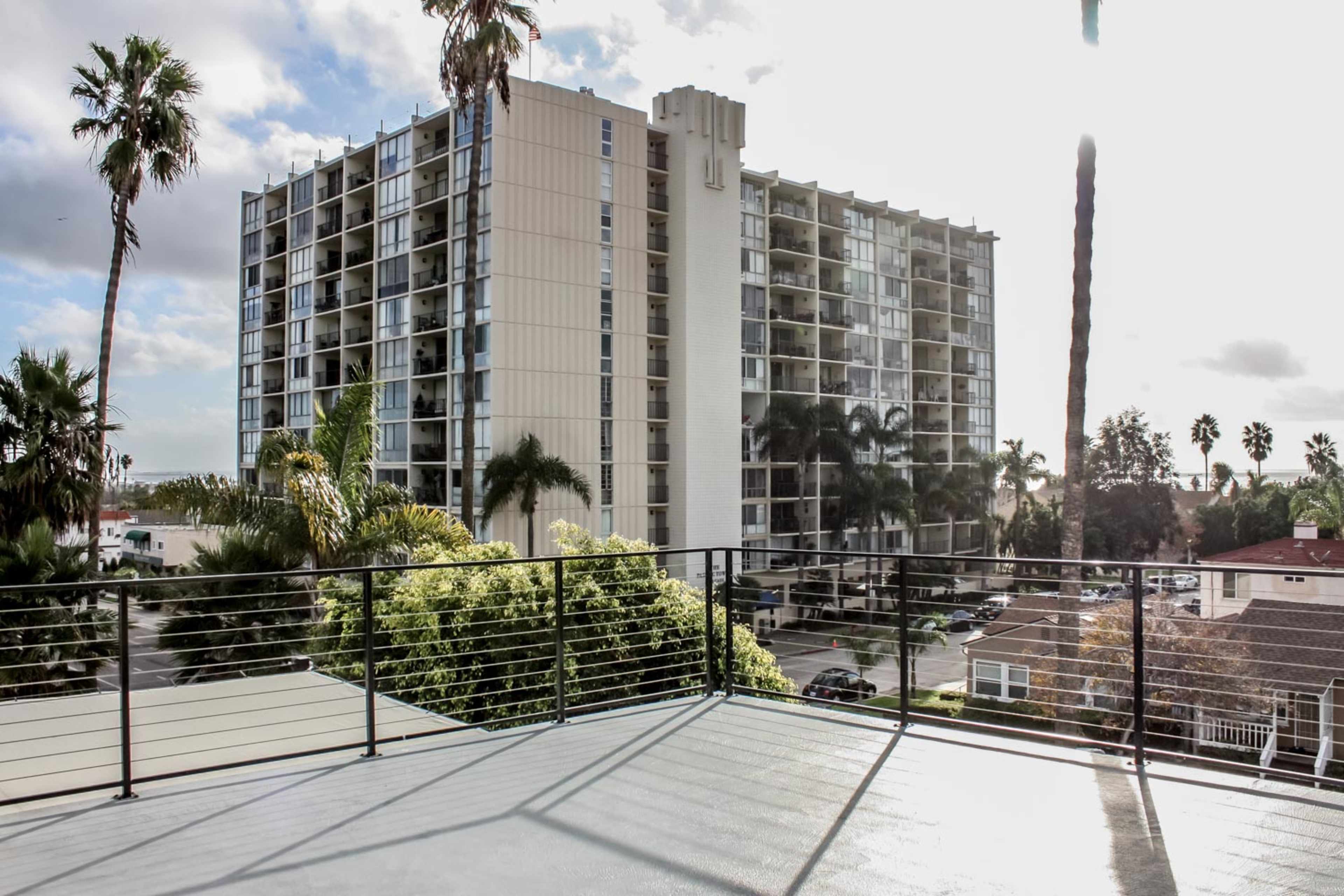 A balcony with a railing overlooks a multi-story apartment building surrounded by palm trees and greenery.