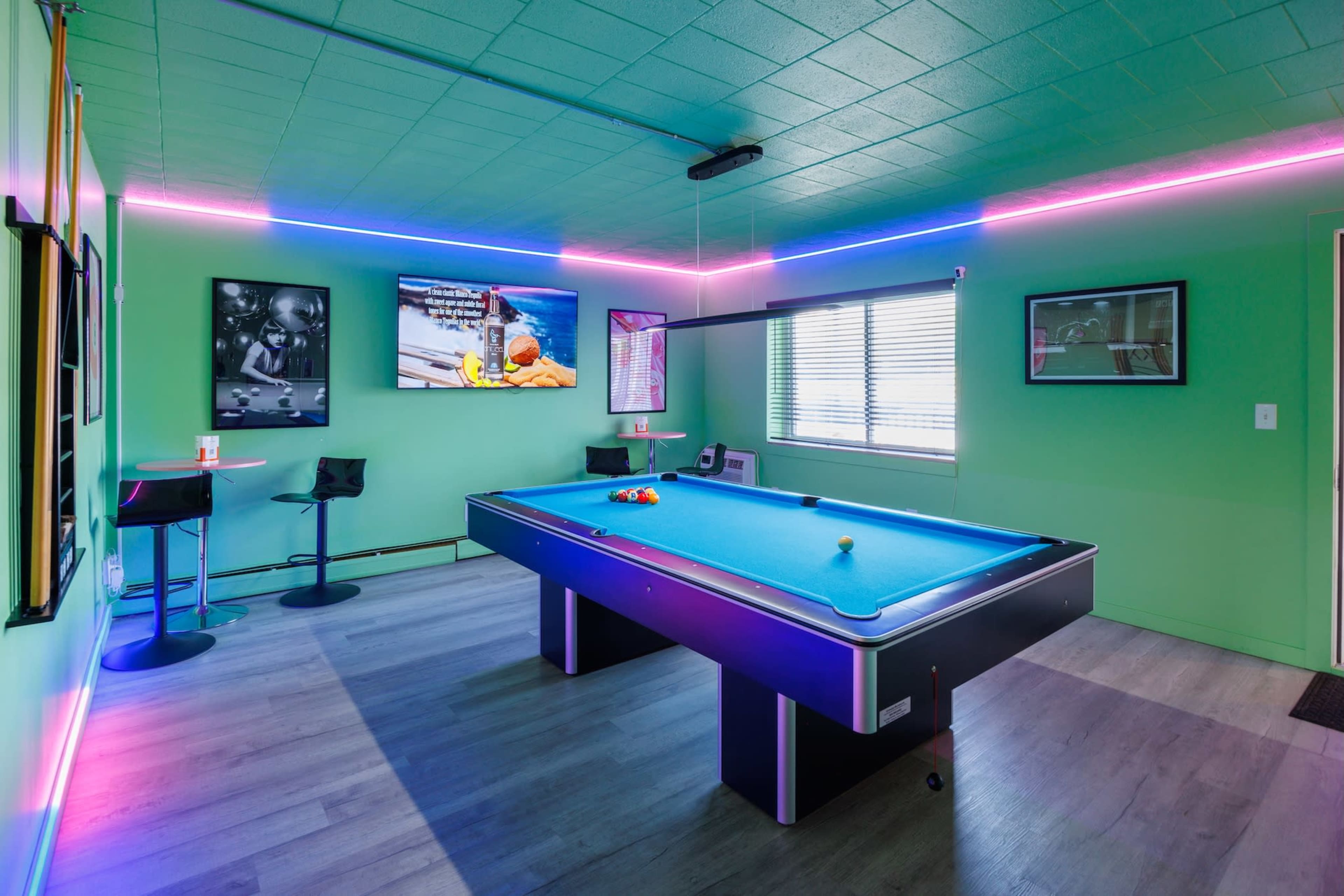 The image shows a brightly colored game room featuring a blue pool table, neon lights, and a wall-mounted television.